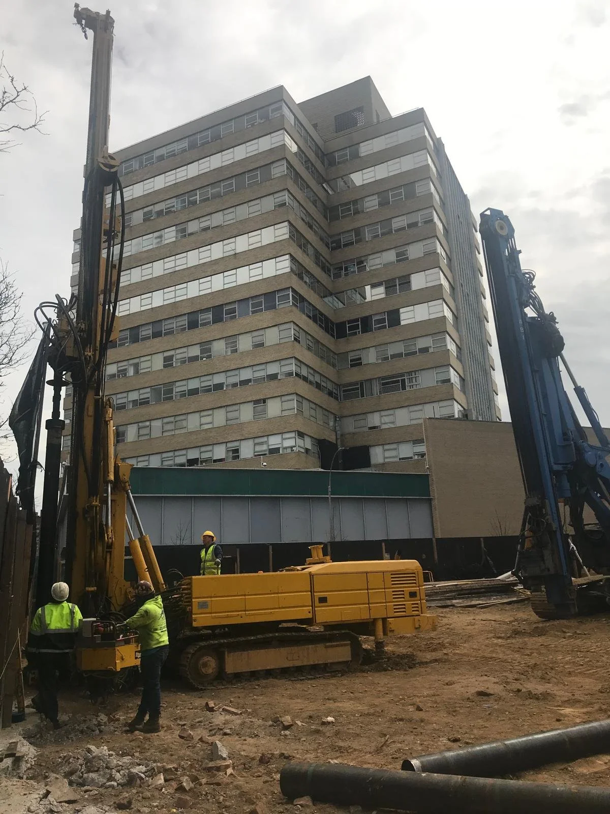 Construction workers in safety gear operating machinery on a construction site with a tall multi-story building in the background.