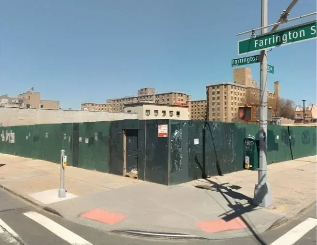 View of a city street corner with a green construction wall, a pedestrian sidewalk, and street signs for Farrington Street and another street. Several buildings are visible in the background under a clear blue sky.
