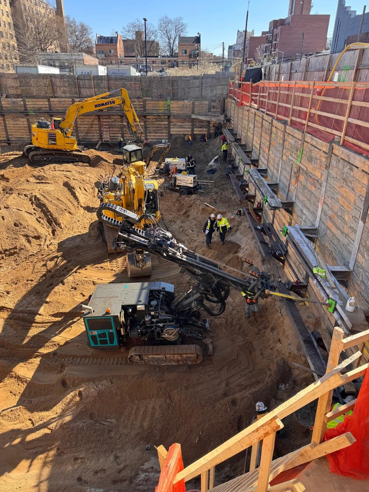 Construction site with excavators and workers digging in a large pit, reinforced by wooden and metal supports, with urban buildings in the background.