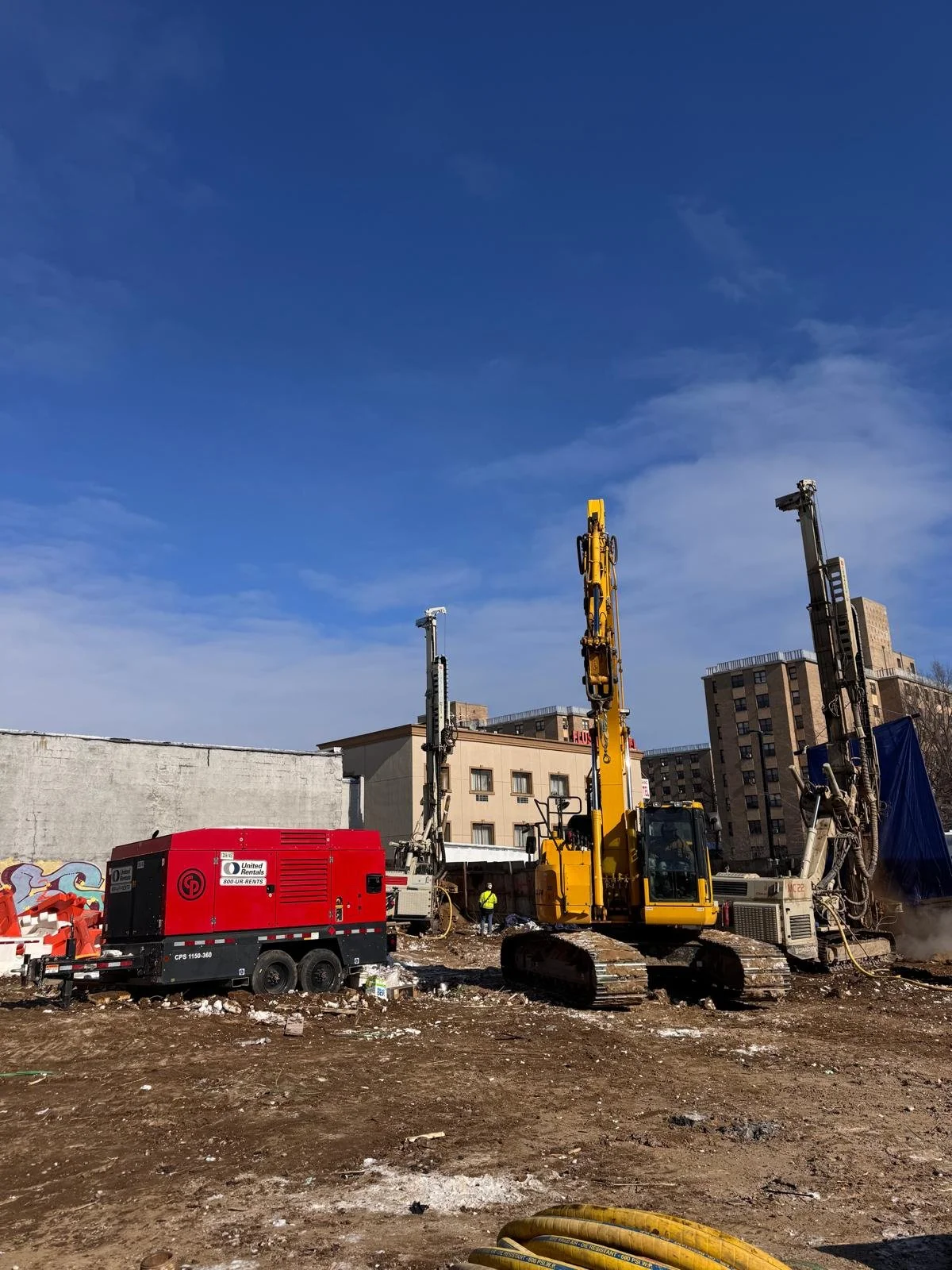 Construction site with heavy machinery including a yellow drilling machine, a red generator, and other equipment on a dirt lot against a blue sky. In the background are apartment buildings and a wall with graffiti.