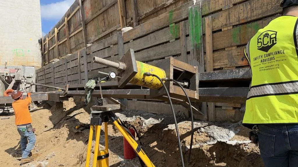 Construction workers, one wearing an orange jacket and helmet, inspecting a construction site with a large steel beam and a hydraulic jack, along with a safety officer wearing a yellow safety vest.