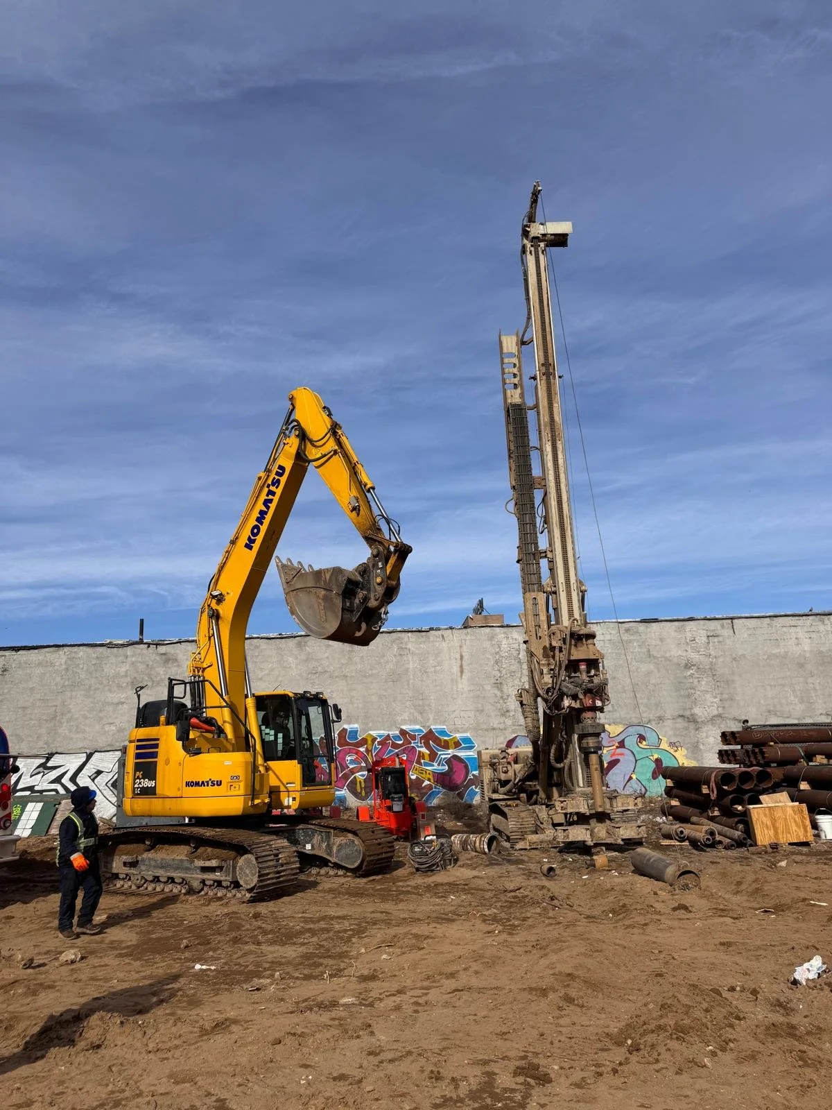 Construction site with a yellow Komatsu excavator and a large drilling machine, workers, and pipes, with graffiti on a concrete wall and a partly cloudy sky.