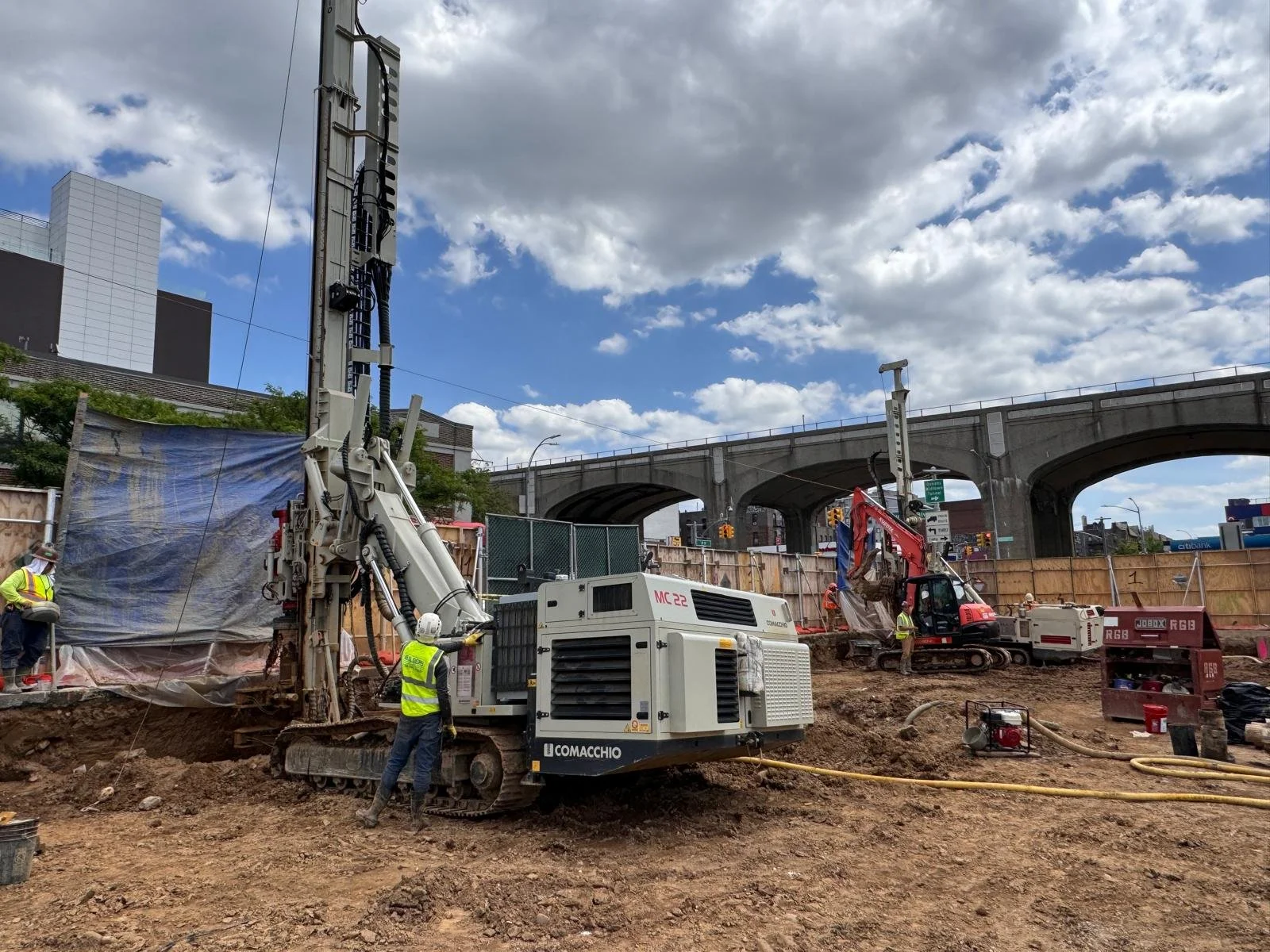 Construction site with heavy machinery, workers in safety vests and helmets, dirt ground, and an overpass in the background under a cloudy sky.