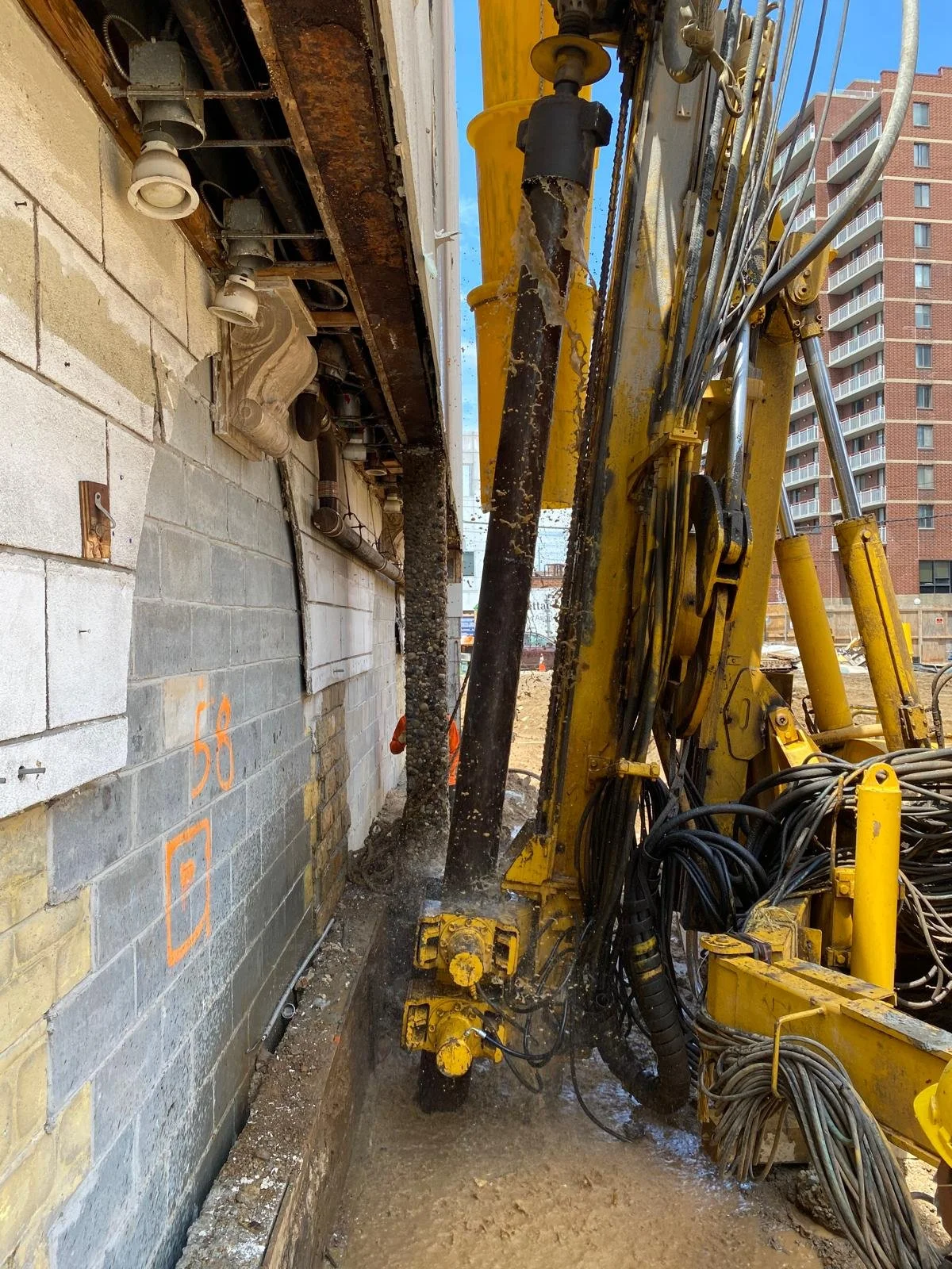 Construction site with a yellow excavator tearing down a building wall, exposing pipes and wiring, with high-rise buildings in the background.
