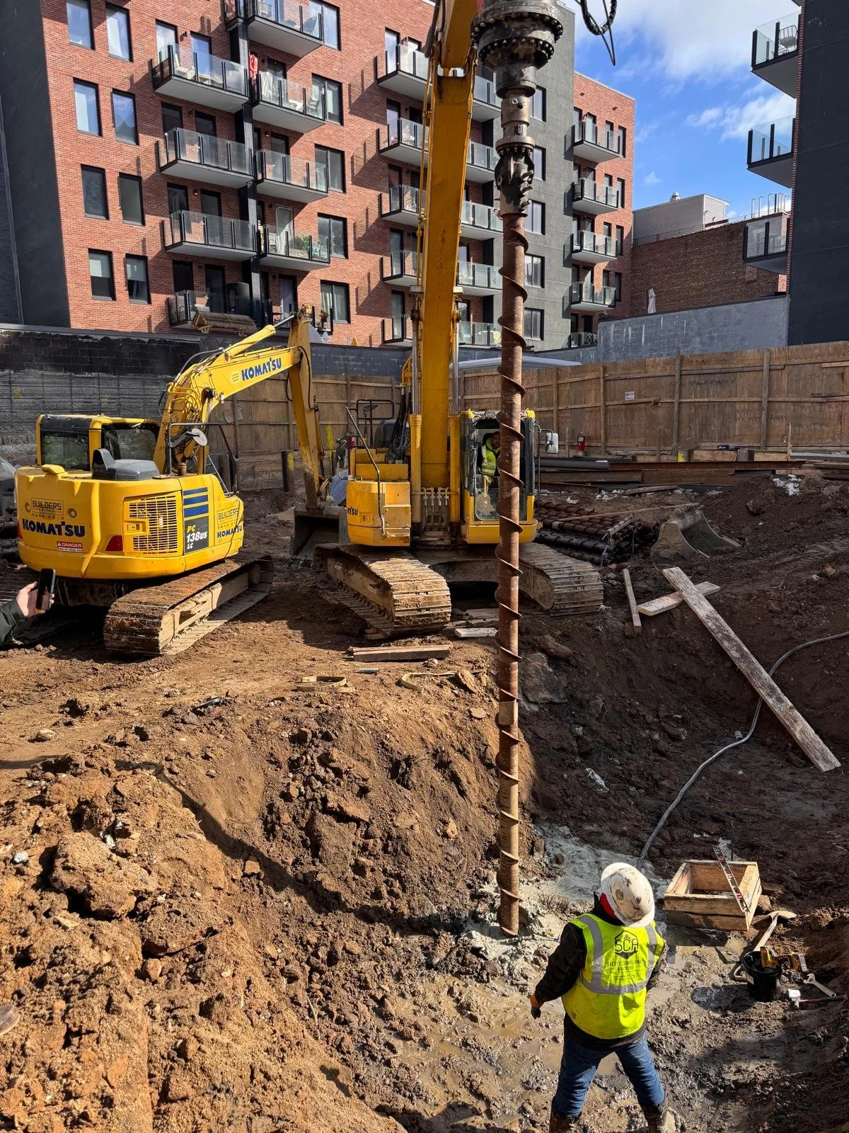 Construction site with large yellow excavators and worker wearing a safety vest and helmet. There is a deep excavation with dirt piled up and building structures in the background.