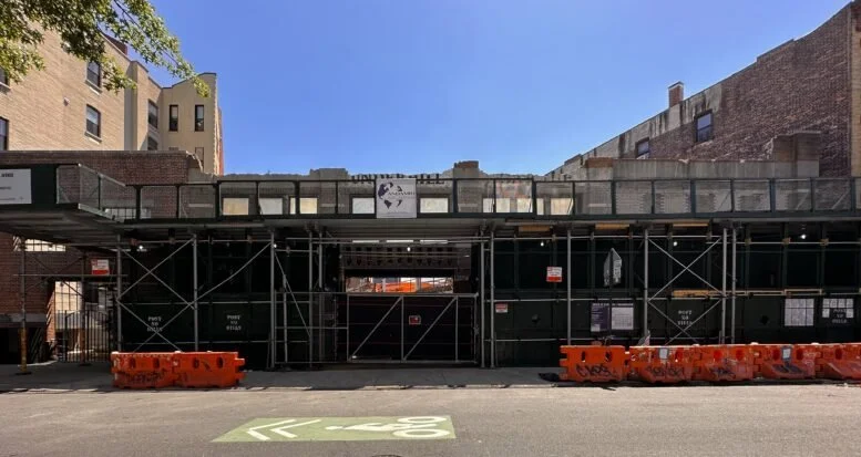 Construction site with black scaffolding and orange barriers in an urban area, brick and concrete buildings in the background, and a blue sky.