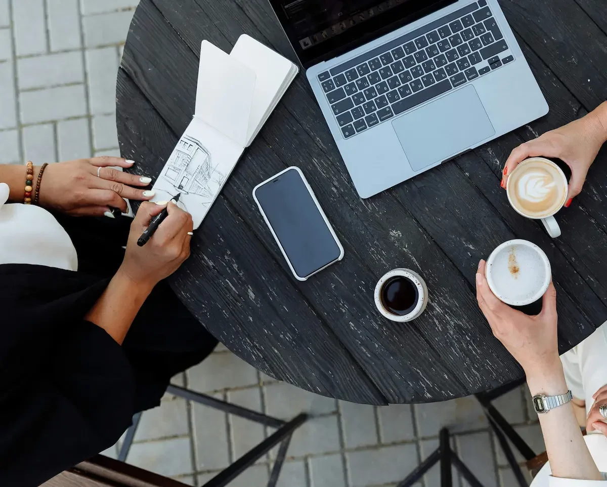 overhead view of three people at table with laptops, a phone, open notebook and cups of coffee.