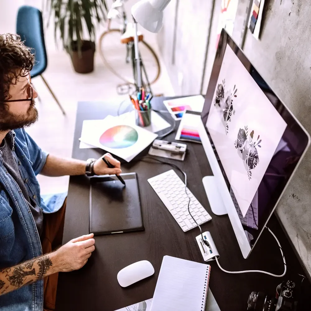 A person working at a desk with a drawing tablet, wacom pen, and a computer monitor displaying graphic designs. The desk also has color swatches, a notepad, a keyboard, a mouse, and various art supplies.