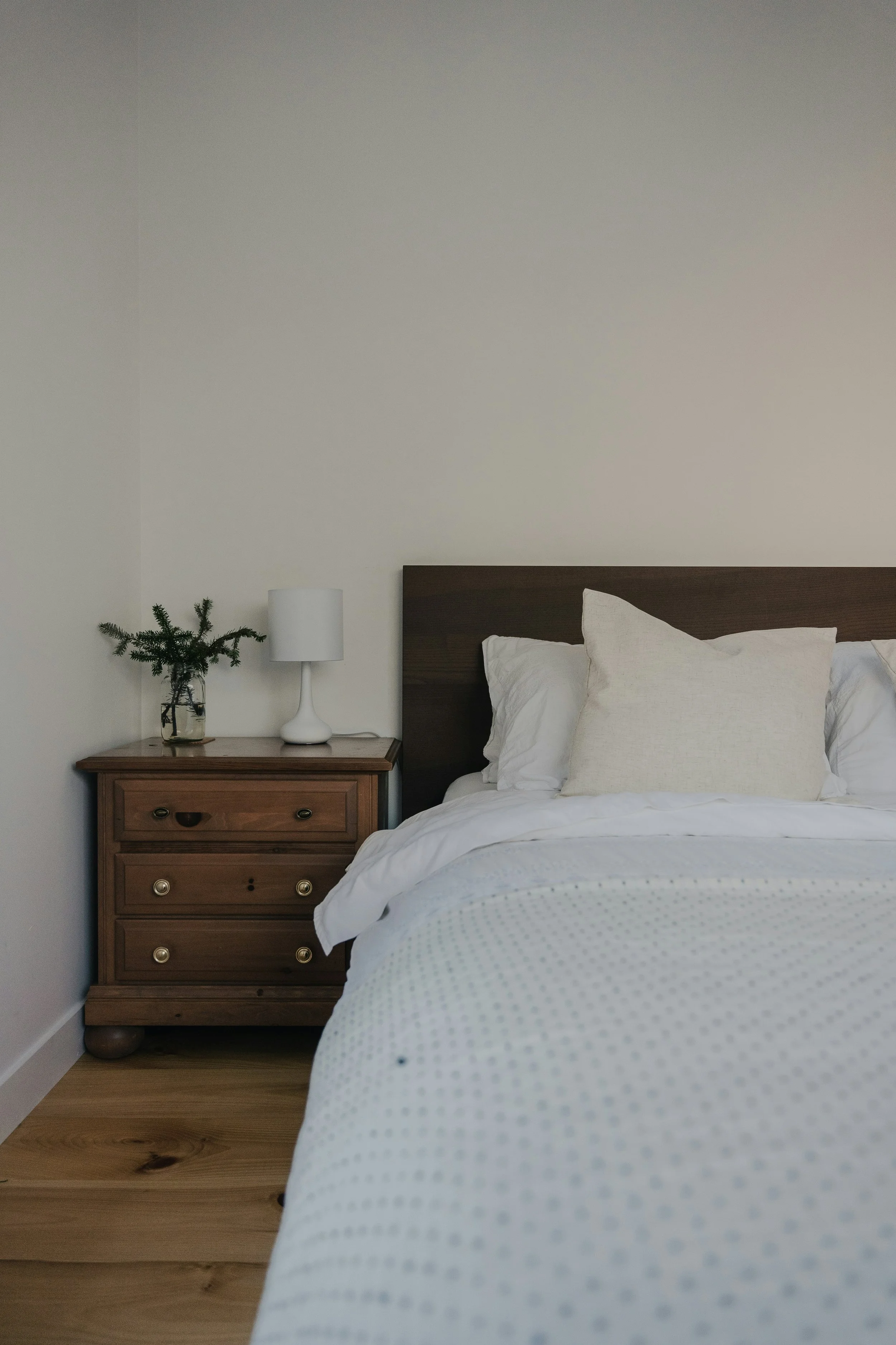 A bedroom featuring a wooden nightstand with brass knobs, a white lamp, a small green plant in a glass jar, and part of a bed with white pillows and a white duvet with tiny black dots. The background is a plain light-colored wall.
