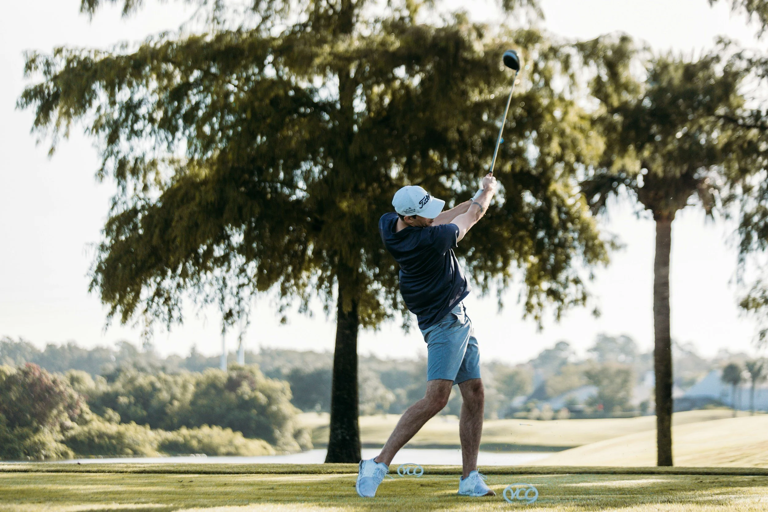 A man practicing golf on a golf course, teeing off with a golf club while standing on a tee box with trees and water in the background.