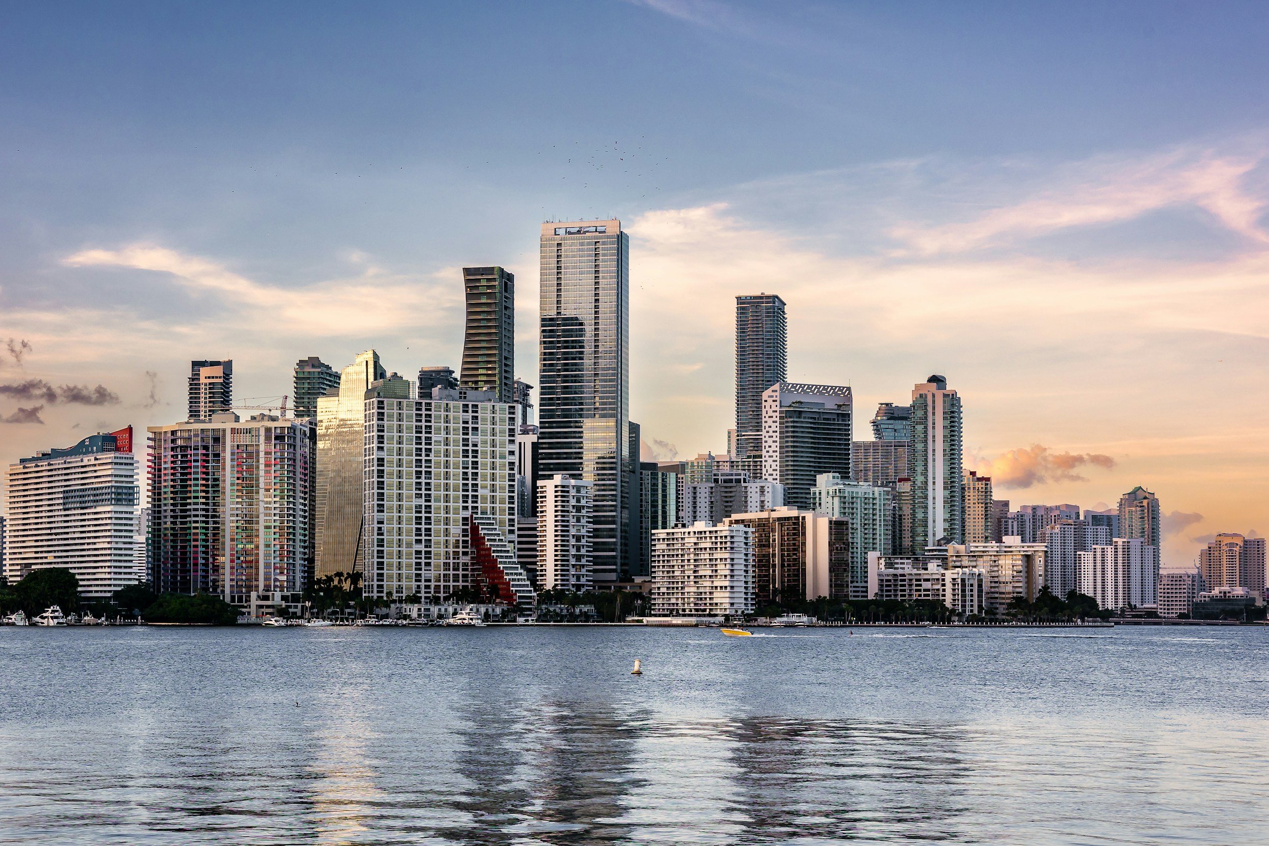 City skyline with tall modern buildings along a waterway at sunset.