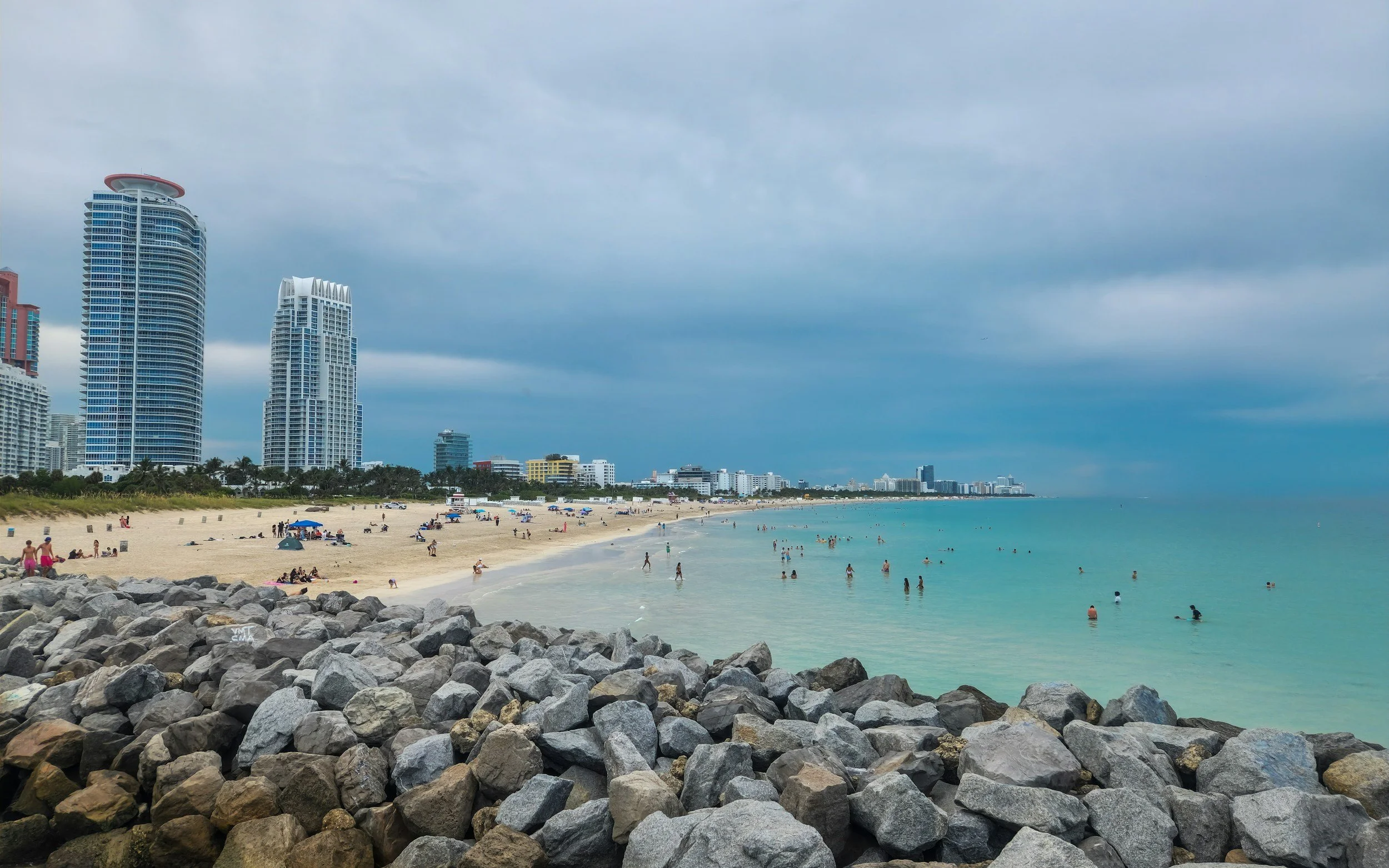 Beach with rocks in foreground, people swimming and relaxing on sandy shore, high-rise buildings along the coast, cloudy sky over ocean.