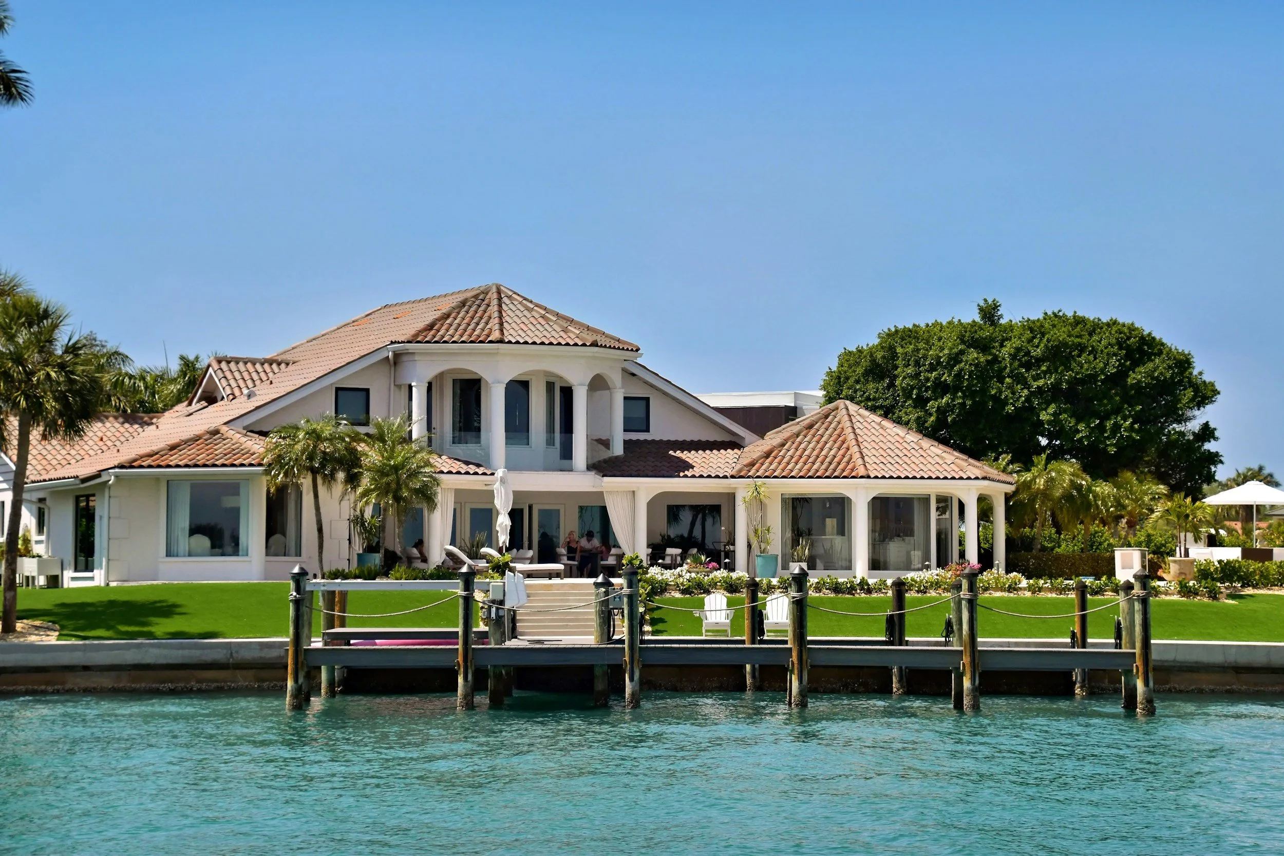 Luxury waterfront house with a tiled roof, palm trees, and a patio, viewed from across the water under a clear blue sky.