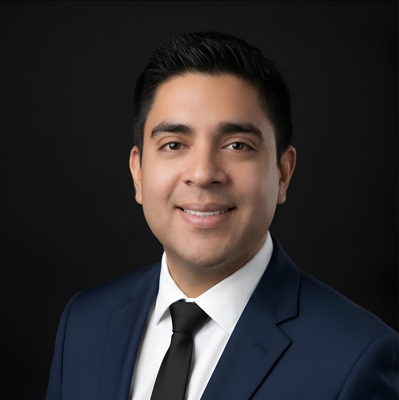 Portrait of a young man wearing a navy suit, white shirt, and black tie, smiling against a black background.