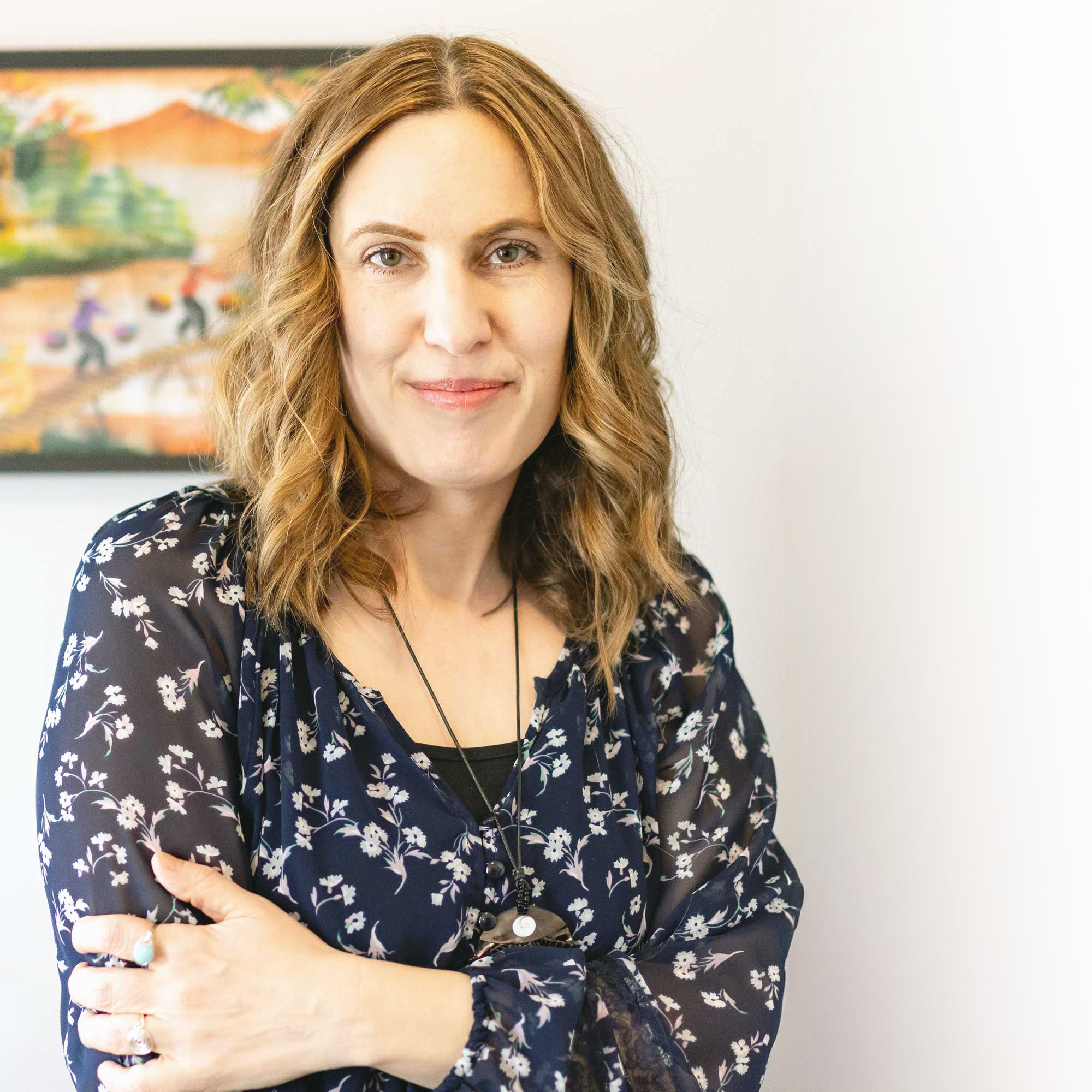 Close-up of a woman with wavy, reddish hair wearing a dark floral blouse, standing against a plain background with a colorful painting visible behind her.