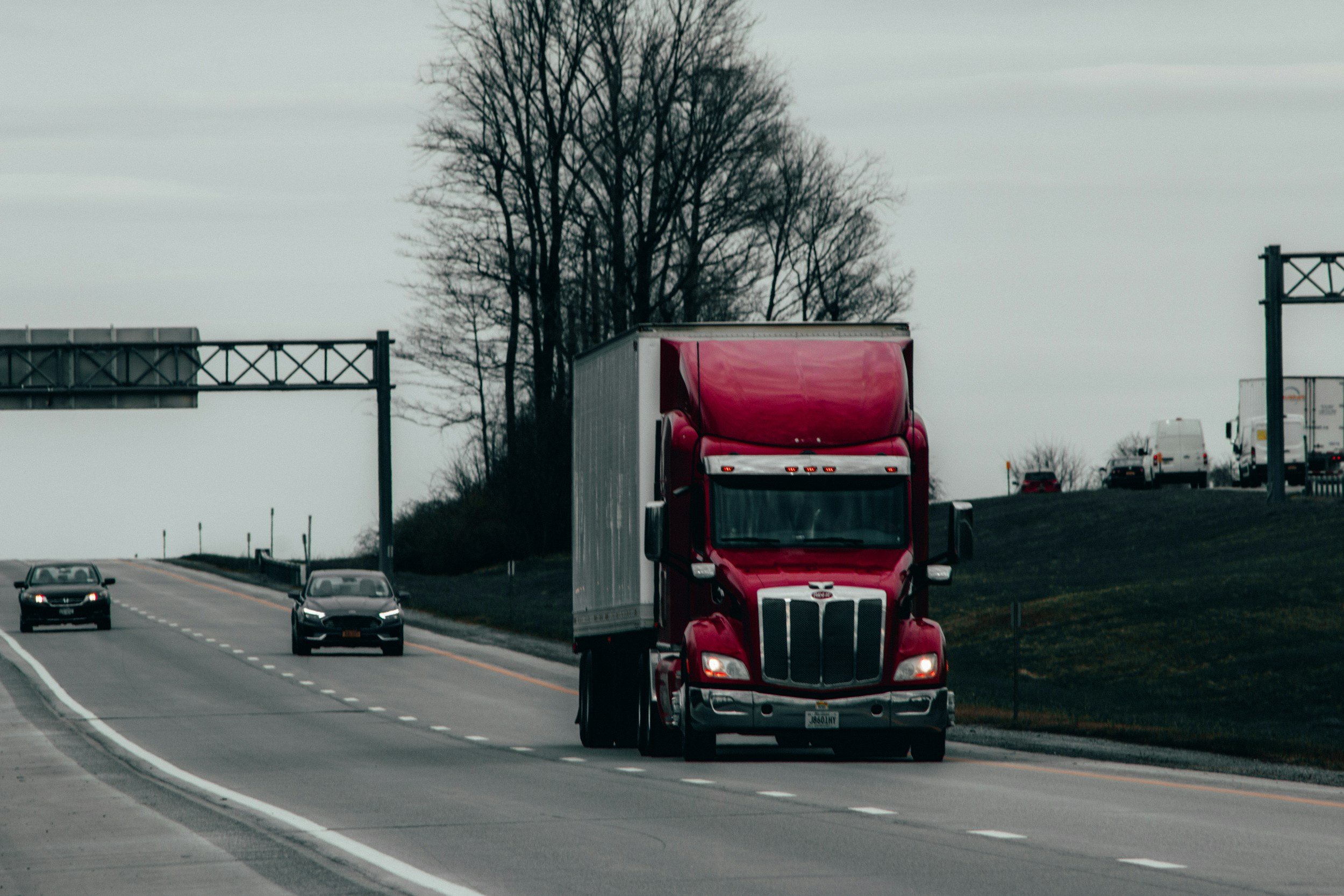 A red semi-truck driving on a highway with other cars and trucks, overcast sky, leafless trees in the background.