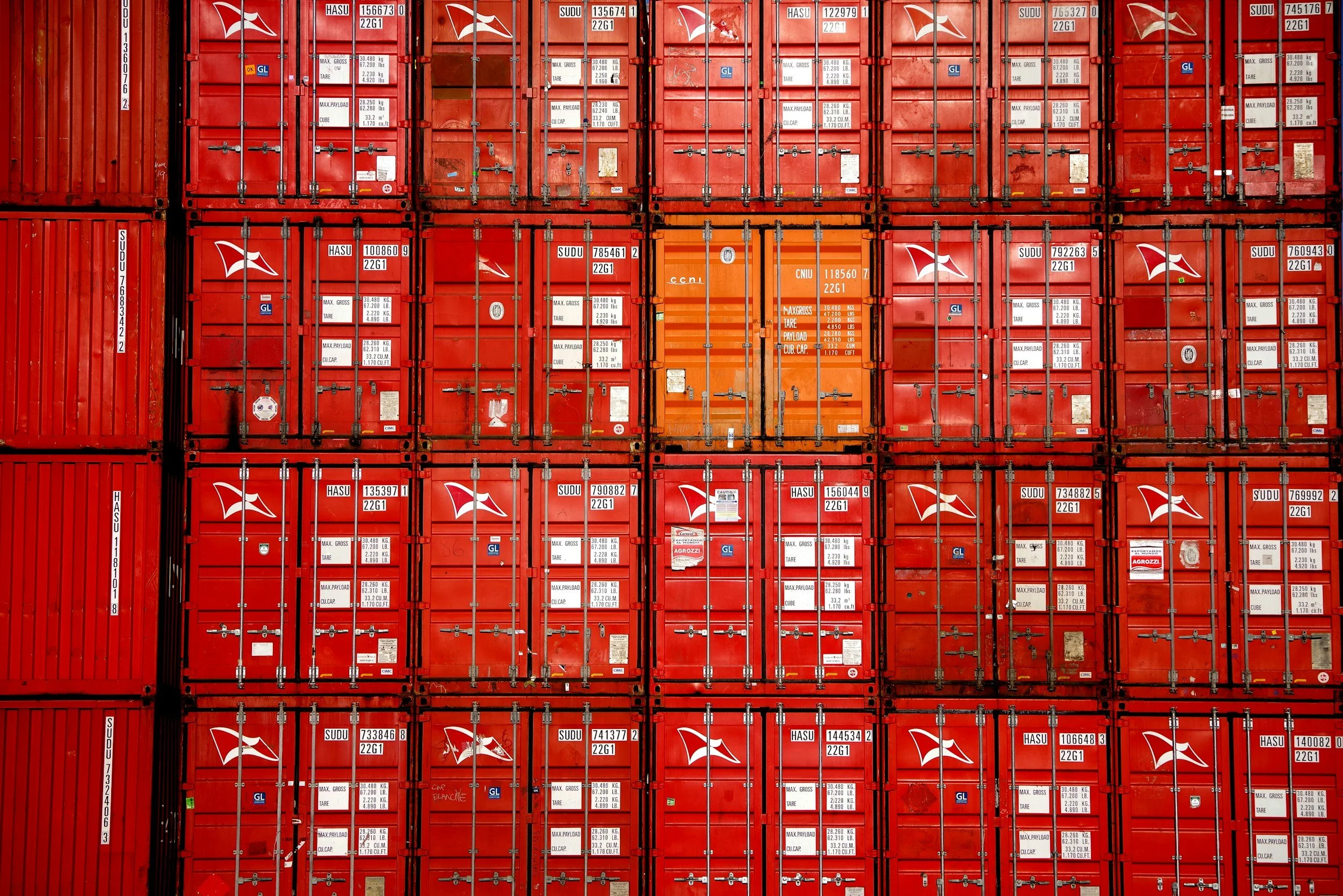 A large number of red shipping containers stacked in a port or freight yard, with various labels, serial numbers, and small logos on each container.