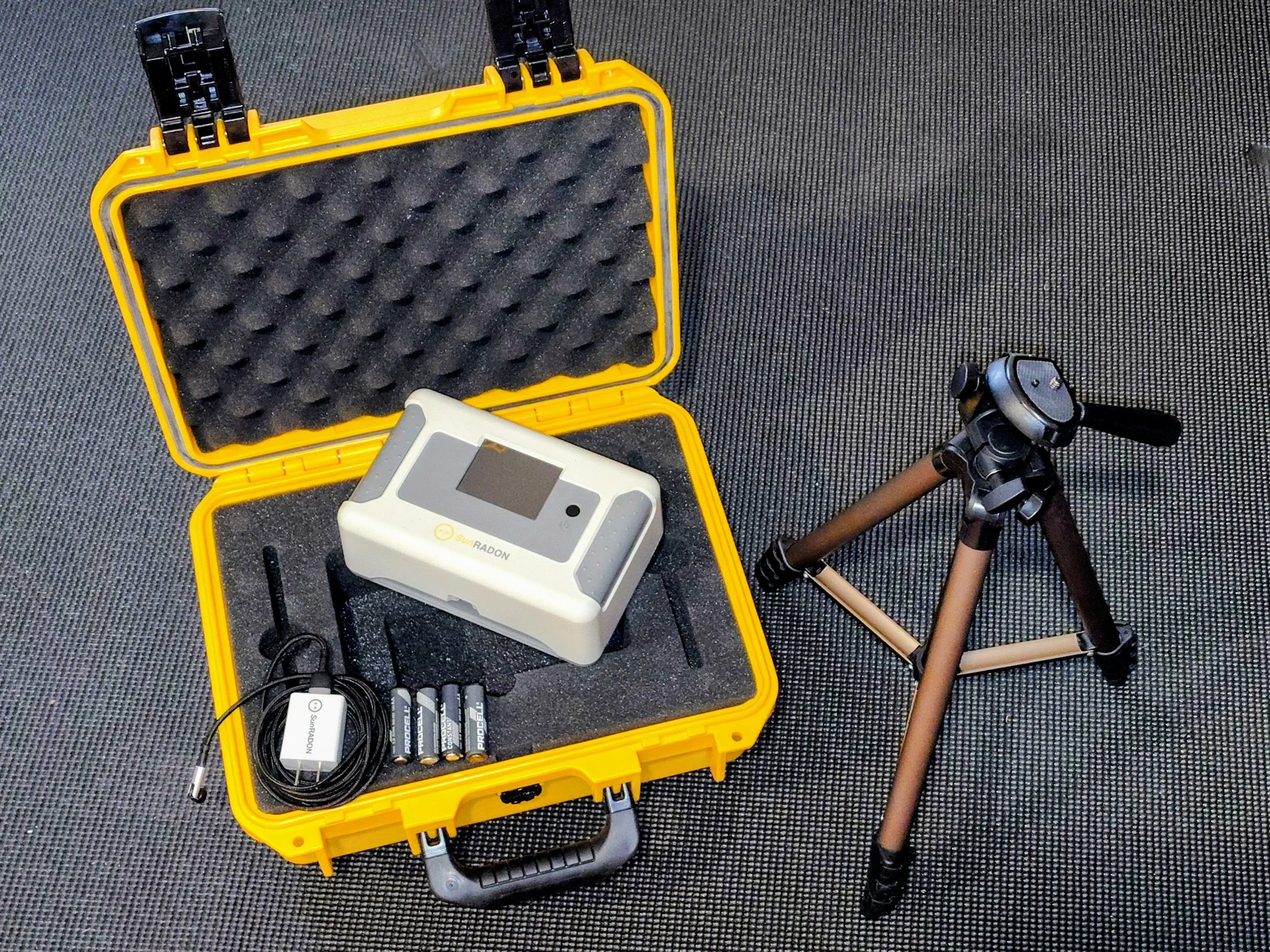 A yellow hard case with foam padding containing scientific equipment including a portable Radon detector, batteries, and other devices, with a tripod and other gear on a gray textured surface.