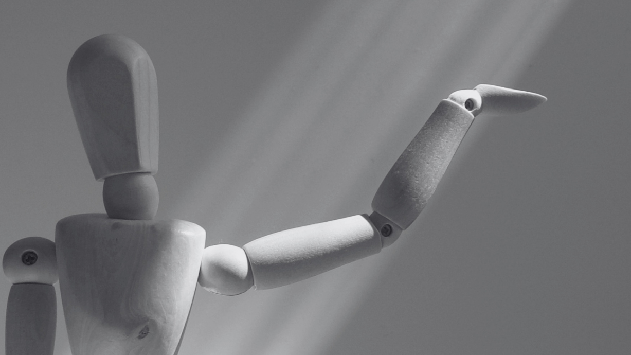 A wooden artist mannequin with one arm extended upward, set against a gray background with light coming from above.