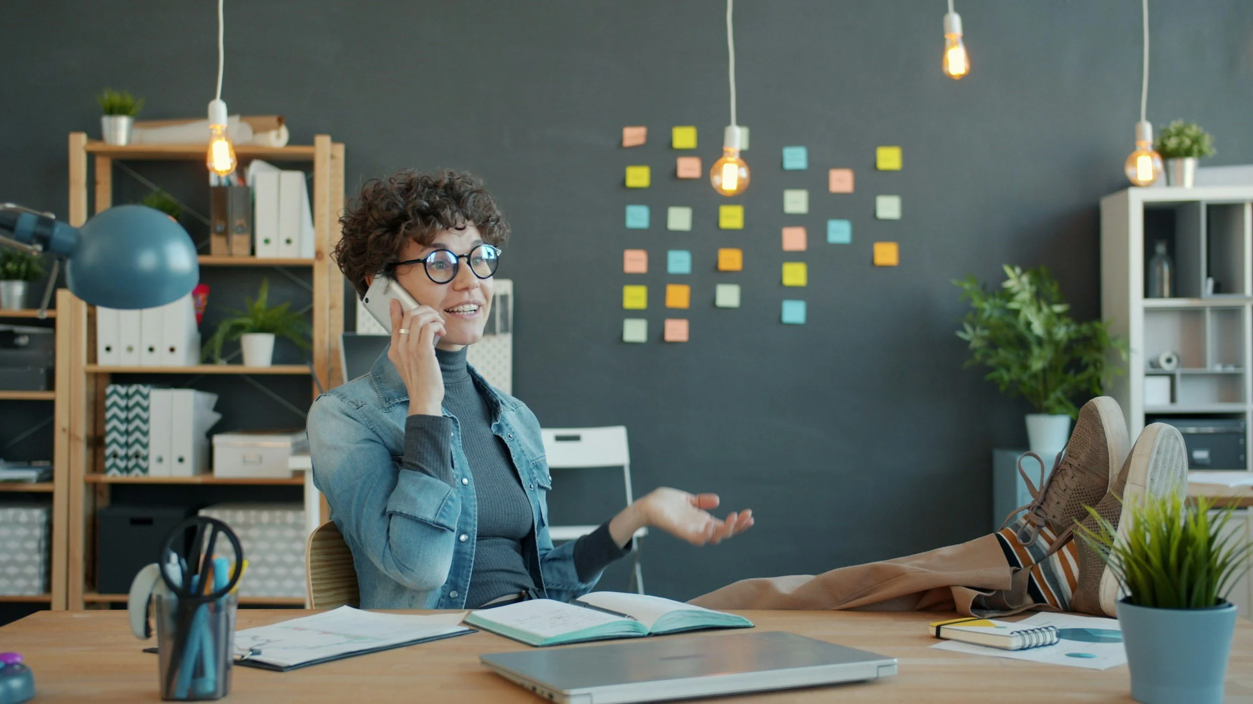 A woman with curly hair and glasses talking on her phone in a modern office, with colorful sticky notes on the wall behind her, a desk with open notebooks, a laptop, and plants.