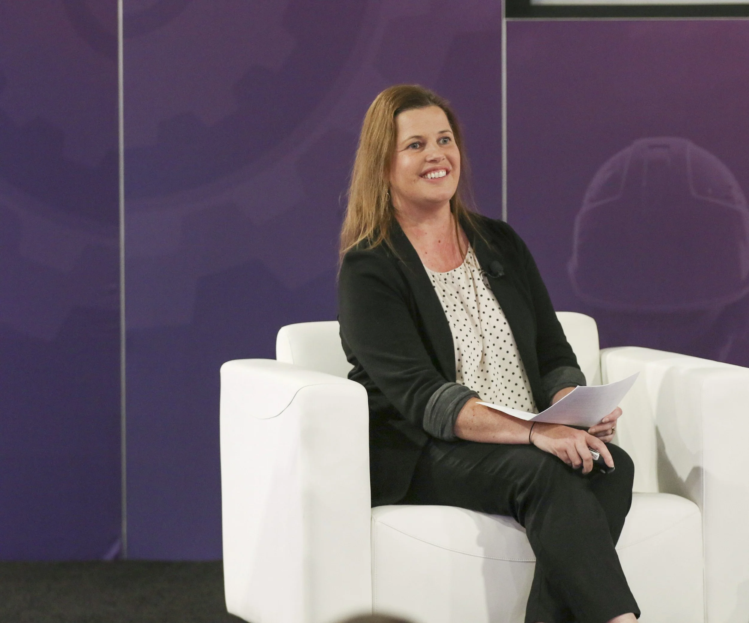 A woman with long brown hair, wearing a black blazer and a white polka-dotted blouse, is sitting on a white chair, smiling, and holding a sheet of paper, with a purple wall with faint rocket illustrations behind her.
