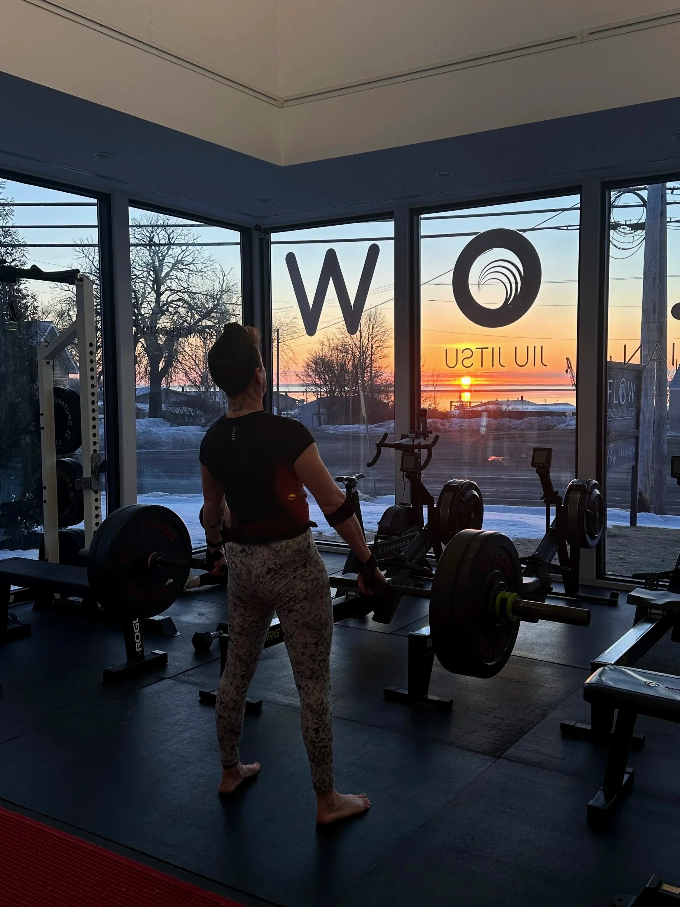 A person with tattoos on their neck and arms is lifting a barbell in a gym, facing large windows showing a sunset over a winter landscape with snow, trees, and power lines.