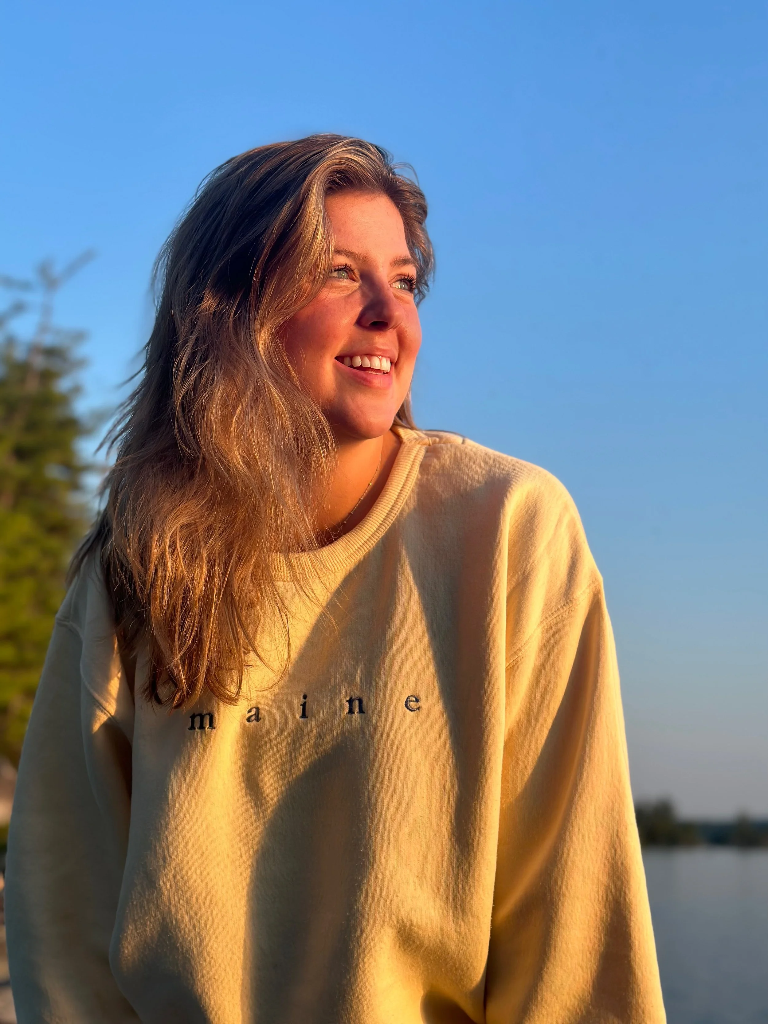 A young woman with long, wavy hair smiling and looking to the side in warm sunlight, wearing a beige sweatshirt with partial text visible, outdoors with a blue sky and trees in the background.