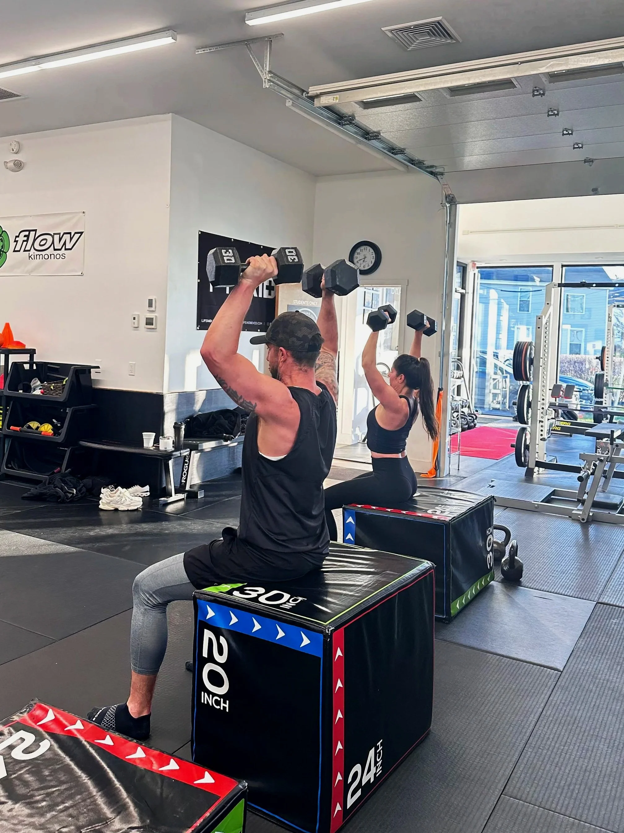People participating in a workout in a gym, lifting dumbbells while seated on bench boxes. The gym has equipment, a wall clock, and a partially open garage-style door letting in sunlight.