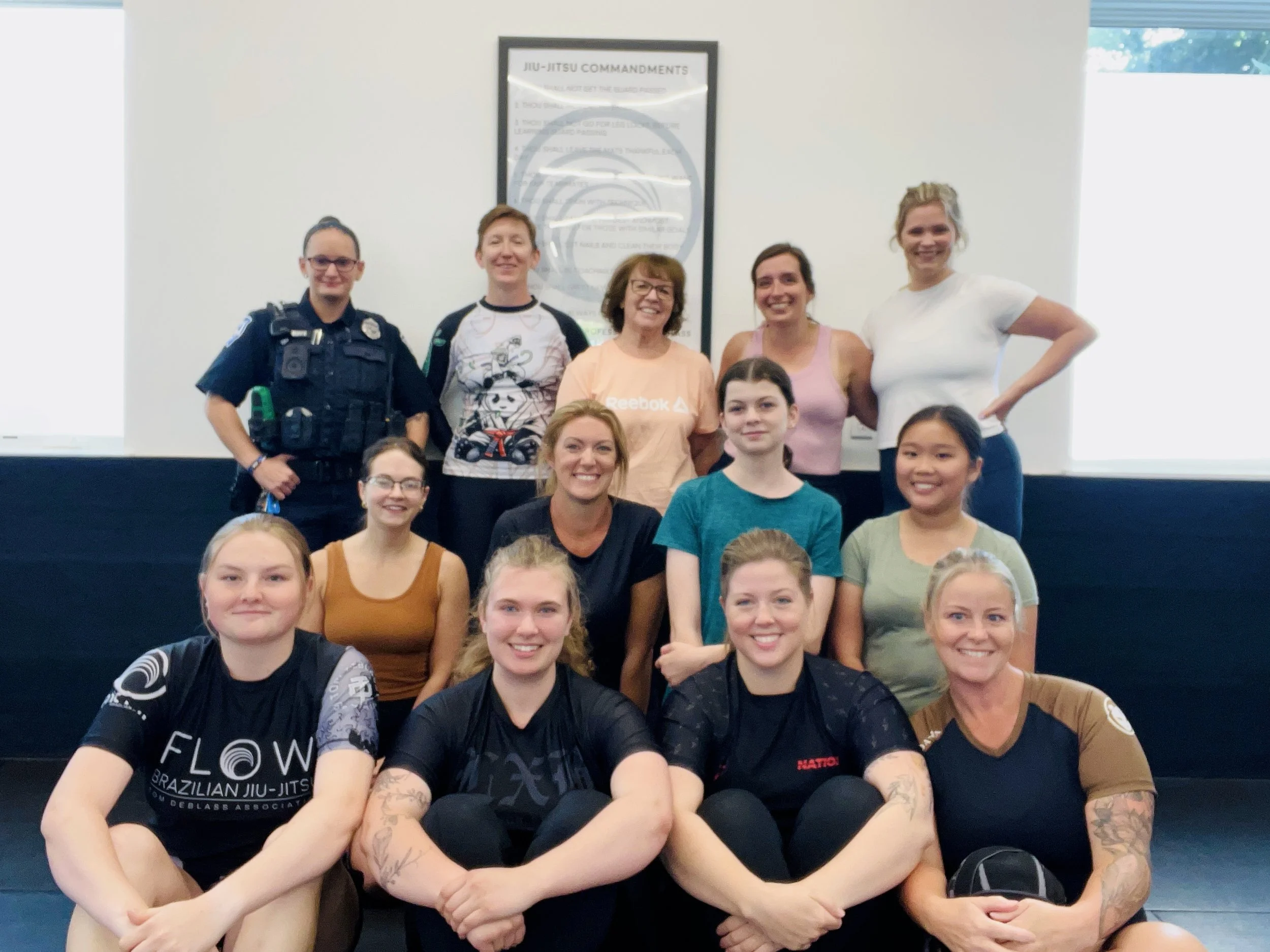 Group of smiling women and girls in a gym or martial arts class, some wearing martial arts uniforms with logos, posing for a group photo in front of a white wall with a Jiu-Jitsu commandments poster.