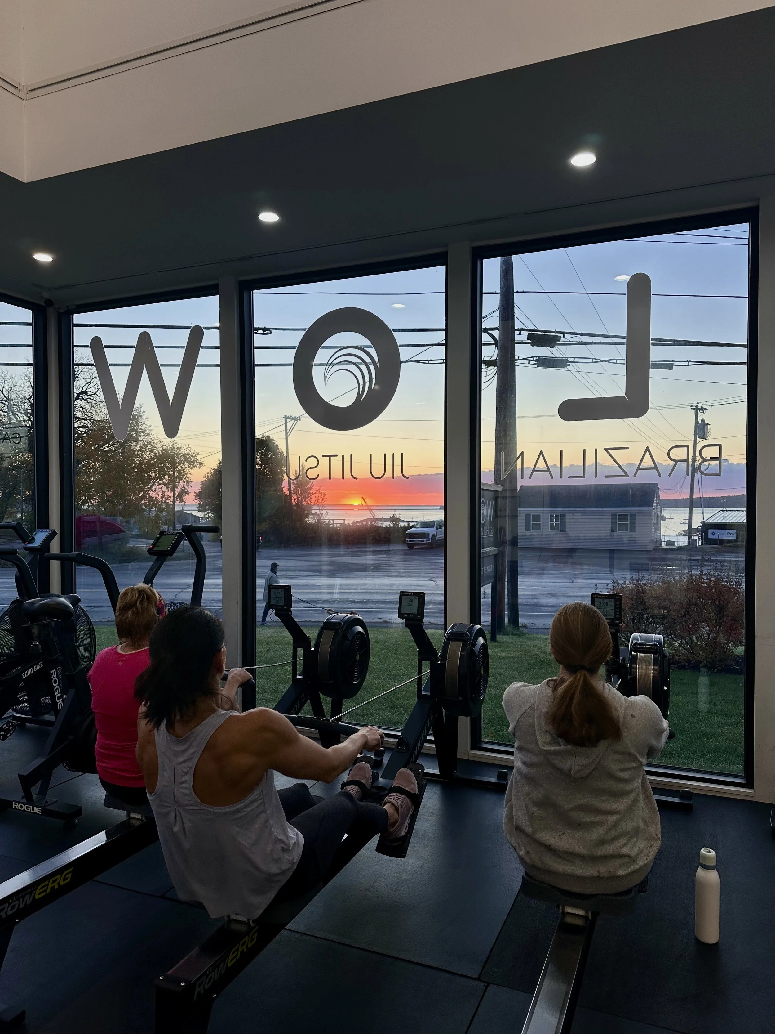 Interior of a gym with three women using rowing machines, looking out of large windows at a sunset over a small town with power lines