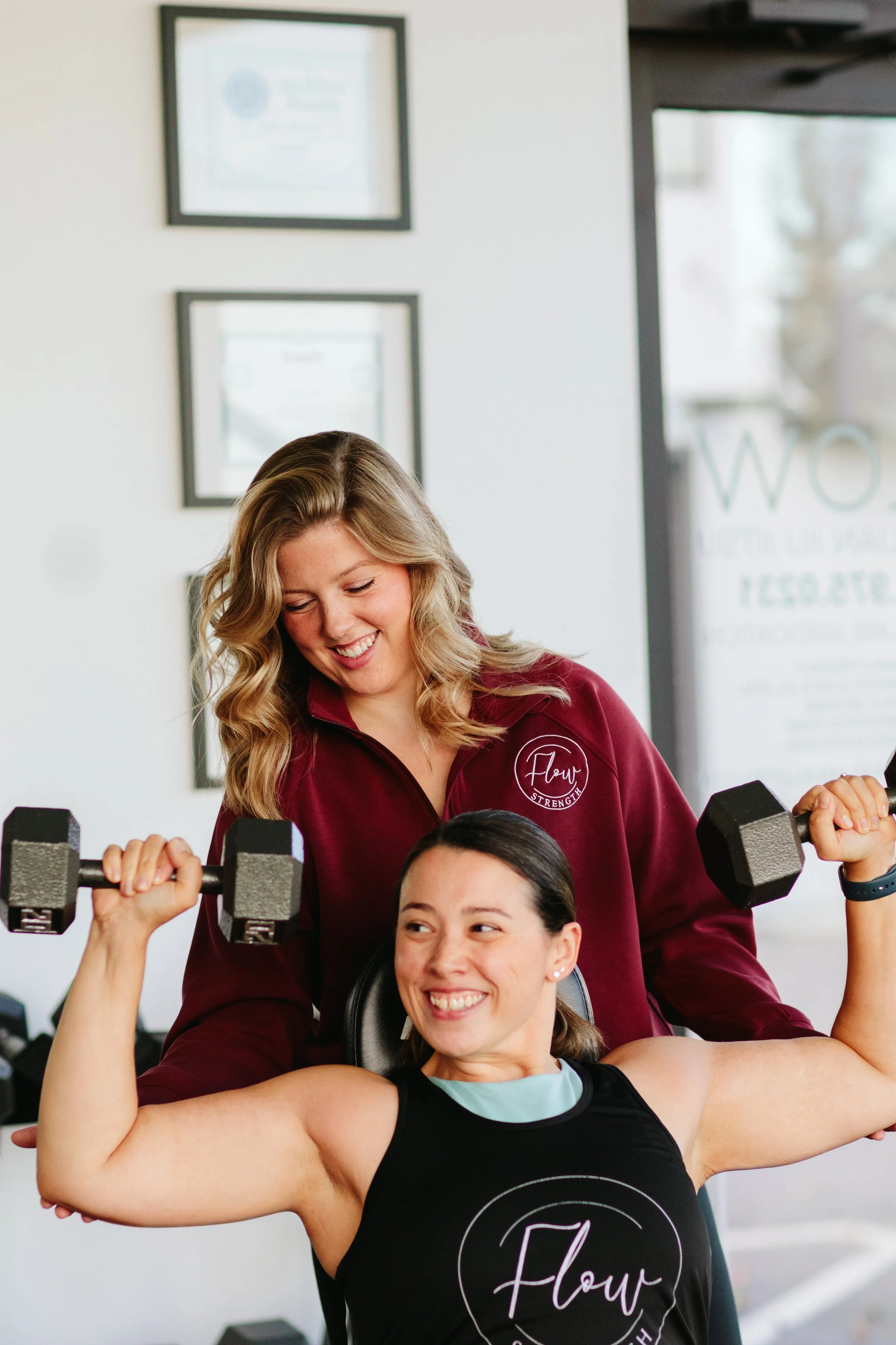 A young woman in a black tank top is lifting dumbbells while smiling and flexing her biceps. A woman in a maroon shirt with a logo that says 'Flour Strength' is standing behind her and smiling, holding a dumbbell, in a gym.