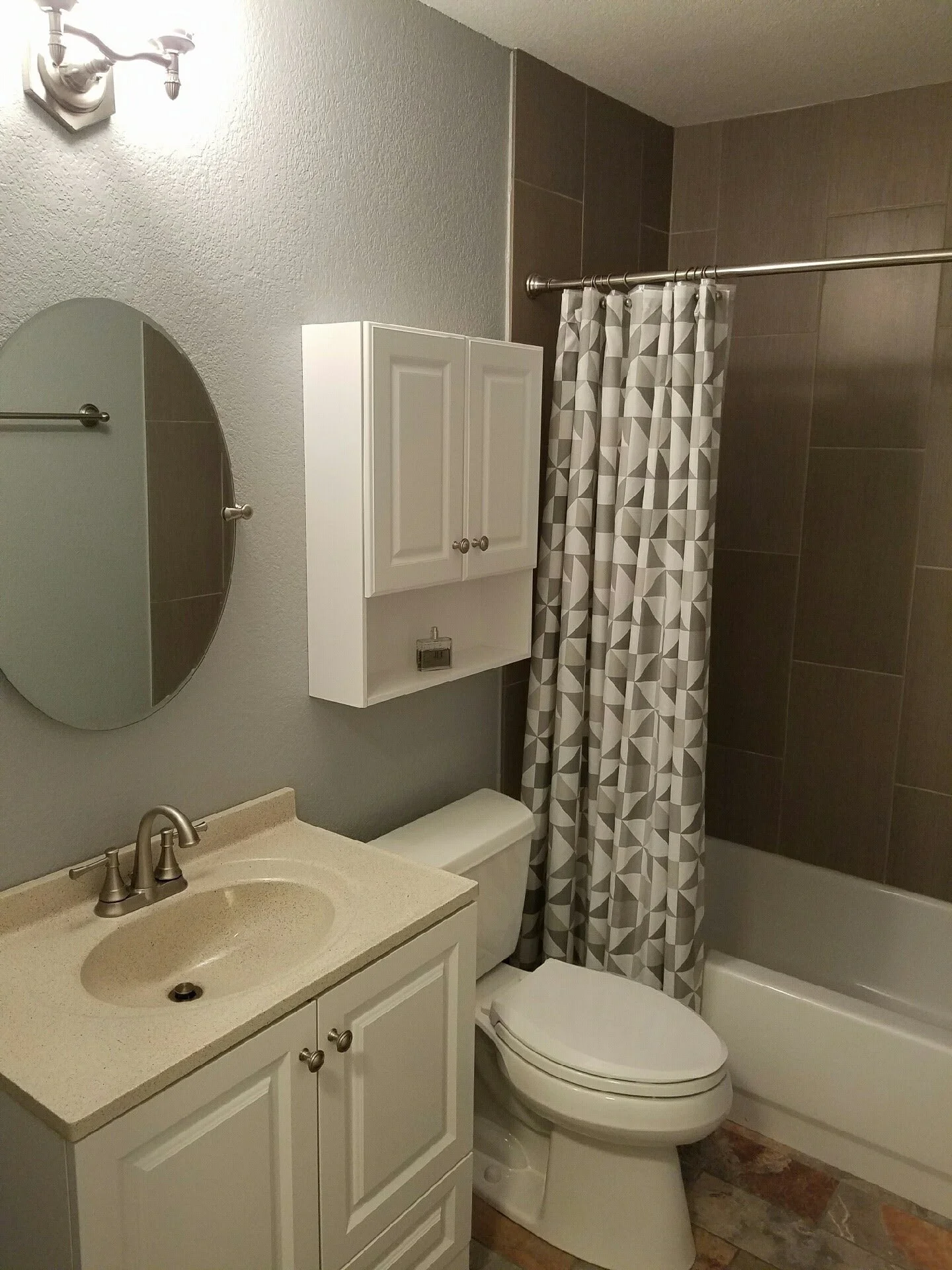 A small bathroom with a beige sink, white cabinets, a mirror, a toilet, a shower with a geometric-patterned curtain, gray tile walls, and a wall-mounted light fixture.