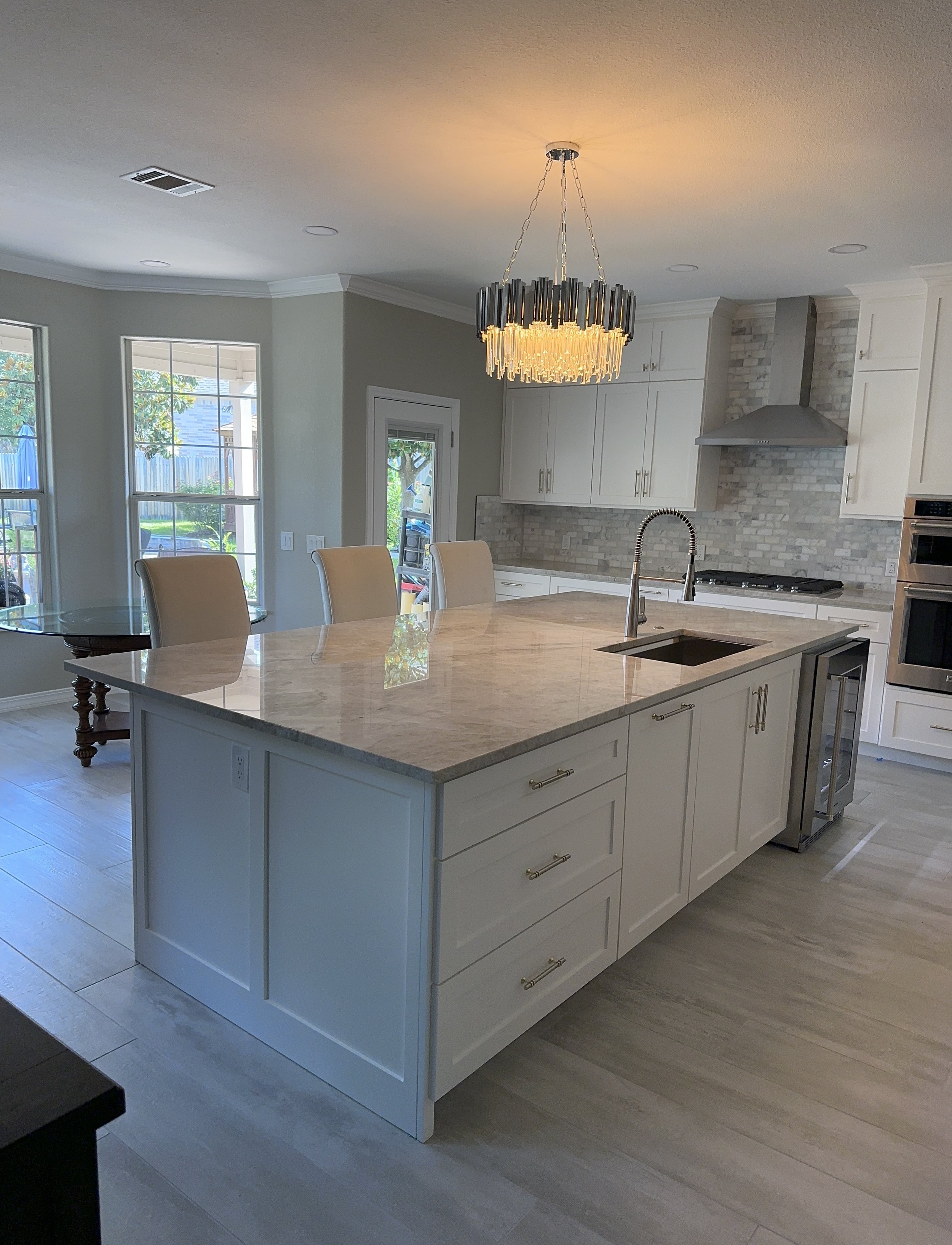 Modern kitchen with a large island, white cabinets, a brick backsplash, a stainless steel range hood, and a chandelier overhead.