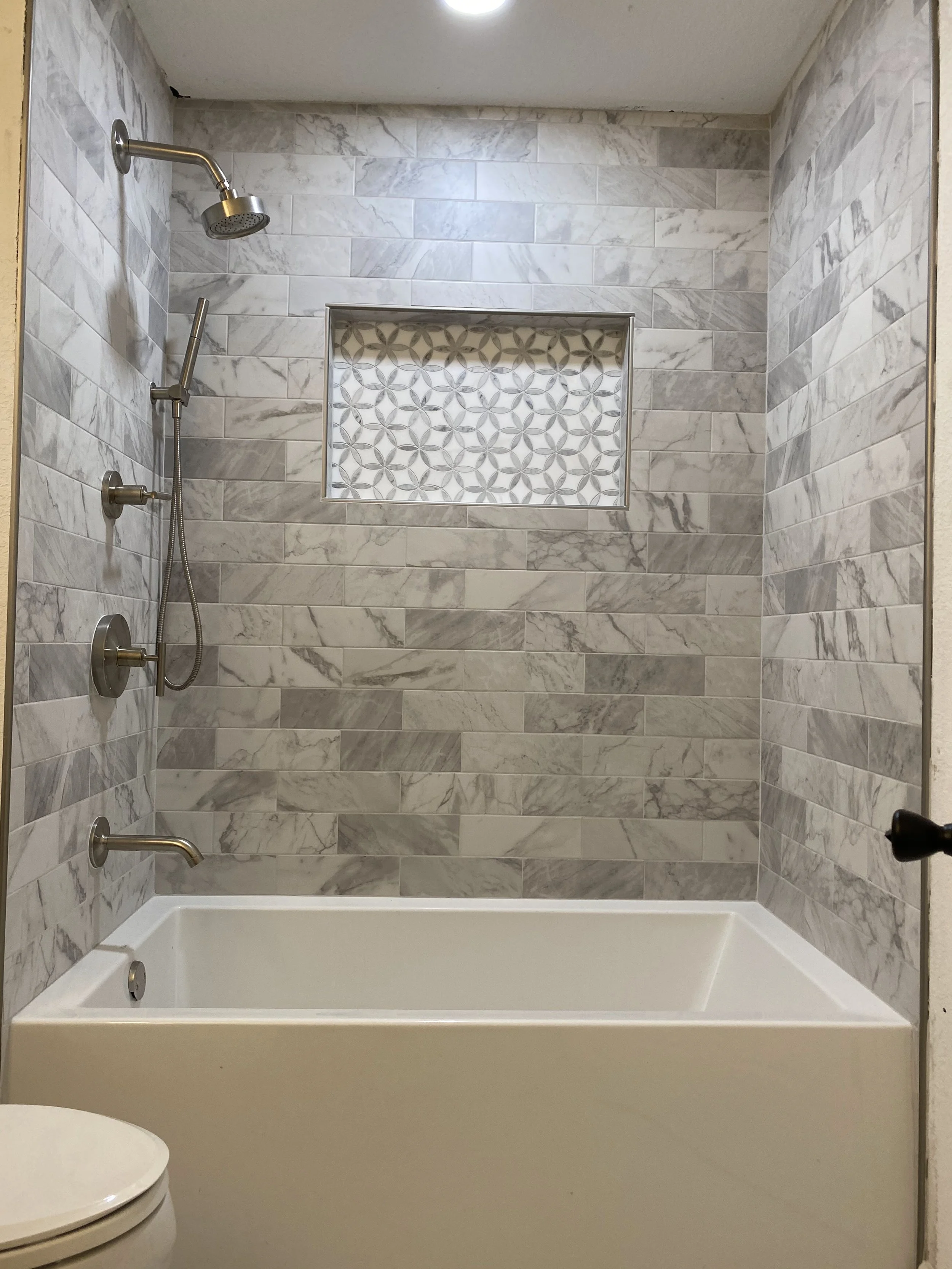 A bathroom shower area with marble tile walls, a built-in niche with decorative tile, multiple shower heads, and a bathtub at the bottom.