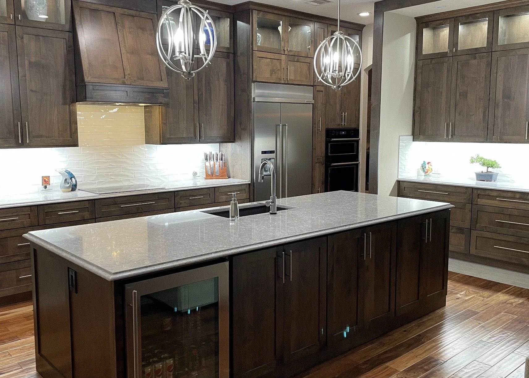 Modern kitchen with dark wood cabinets, white subway tile backsplash, central island with white countertop, stainless steel refrigerator, double oven, and decorative pendant lighting fixtures.