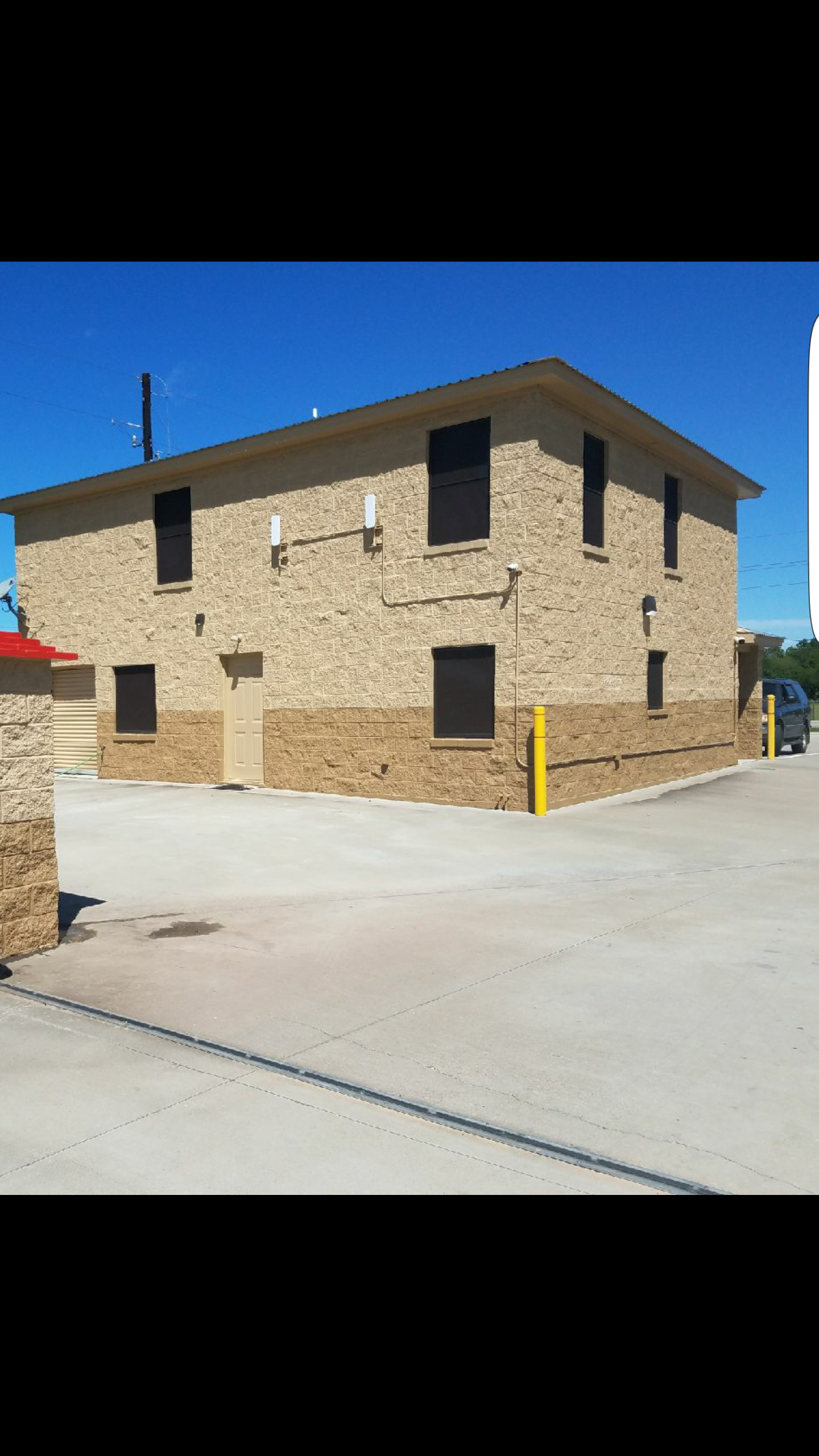A two-story beige brick building with six dark windows, a closed beige door, a yellow pole, and a parking area under a clear blue sky.