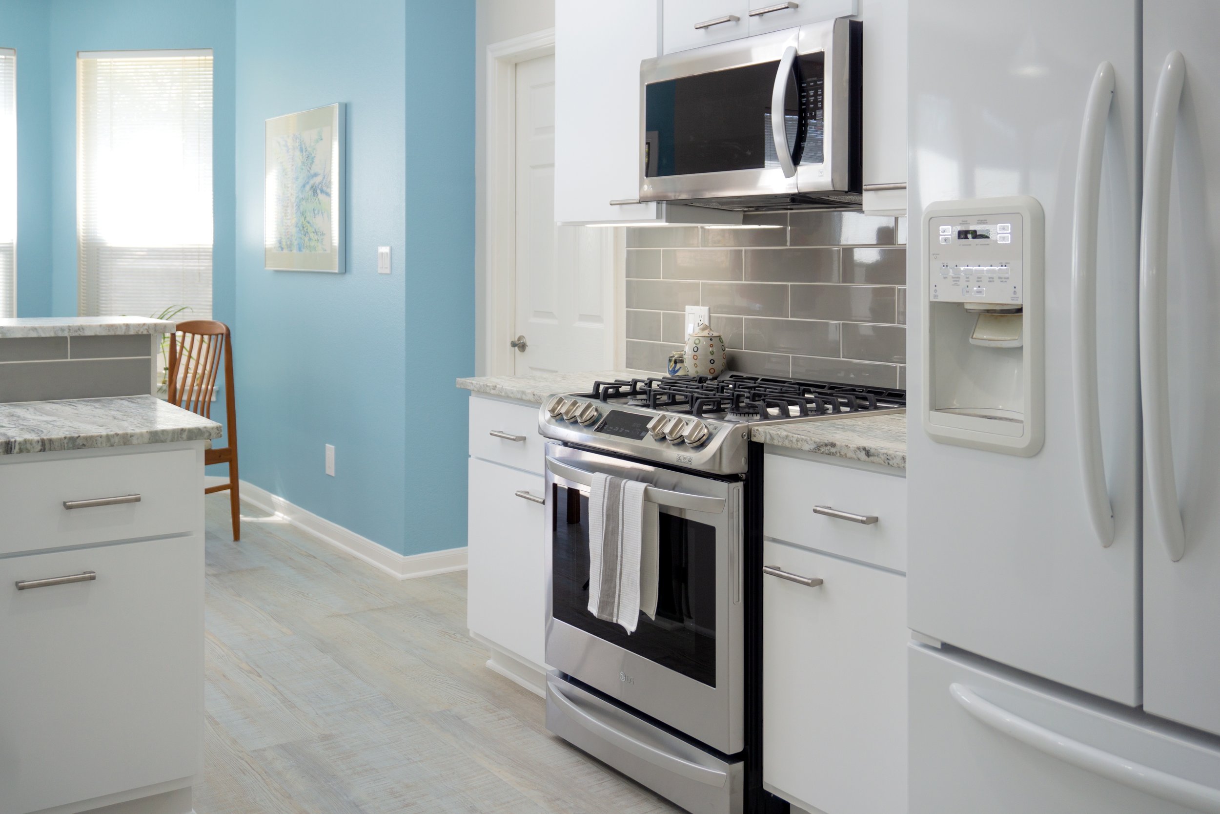A modern kitchen with light blue walls, white cabinetry, stainless steel microwave, gas stove, and a white refrigerator. There is a small countertop area with a decorative jar, a dining table with a wooden chair, and a window with blinds.