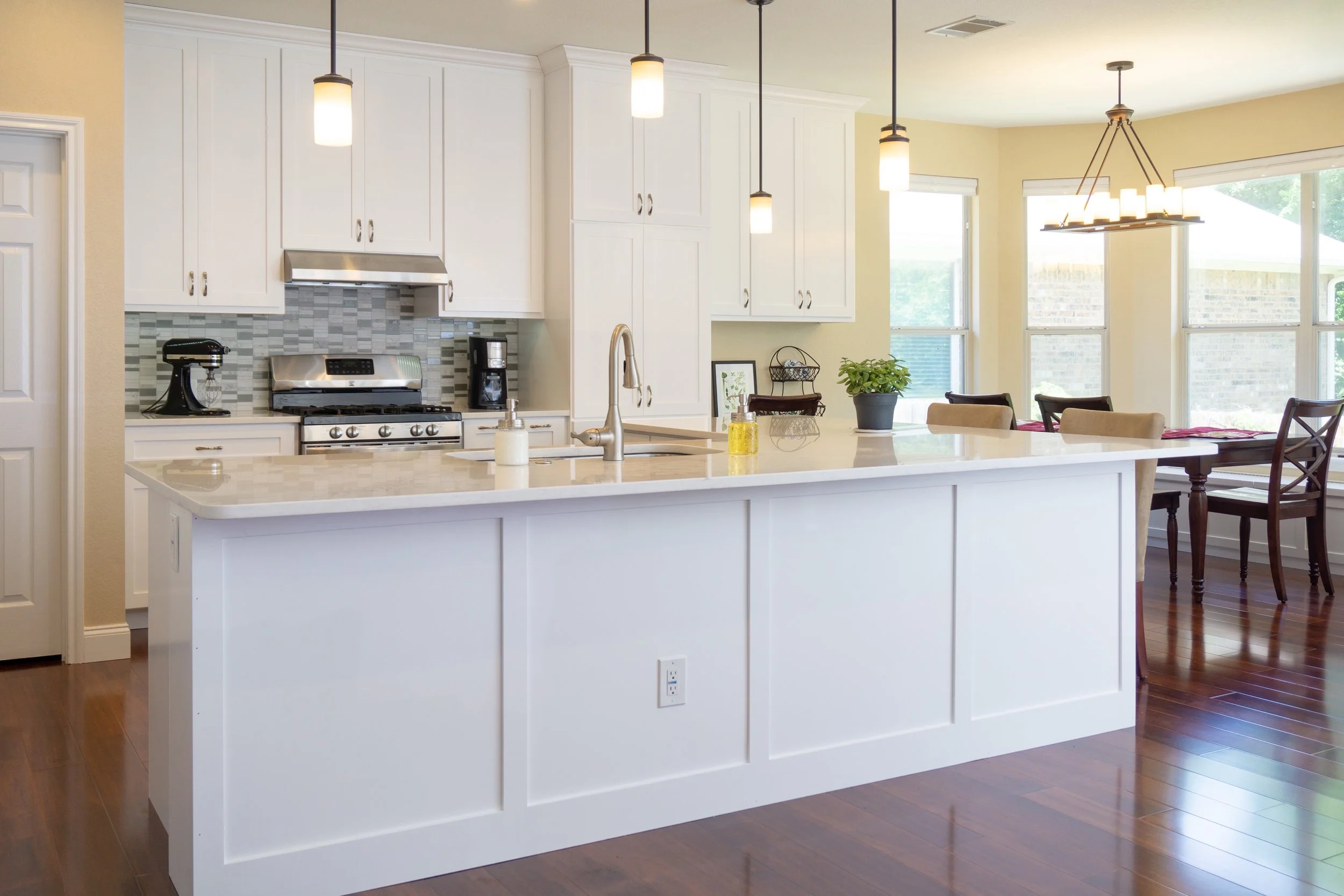 Modern kitchen with white cabinets, a kitchen island, pendant lighting, hardwood flooring, and a dining area with large windows and a chandelier.