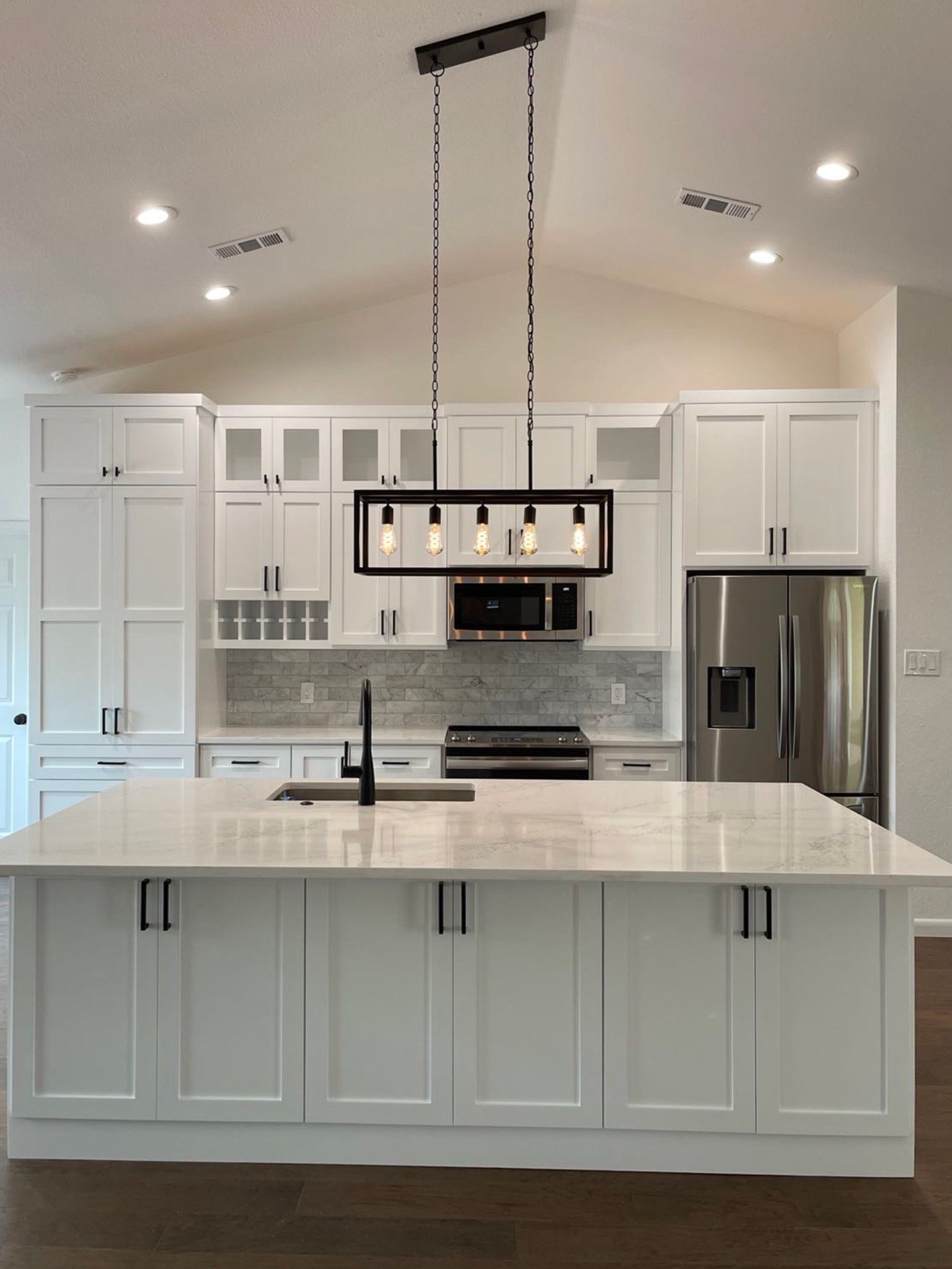 Modern kitchen with white cabinets, a large white island, stainless steel refrigerator, microwave, black light fixtures with exposed bulbs, and a brick-patterned backsplash.