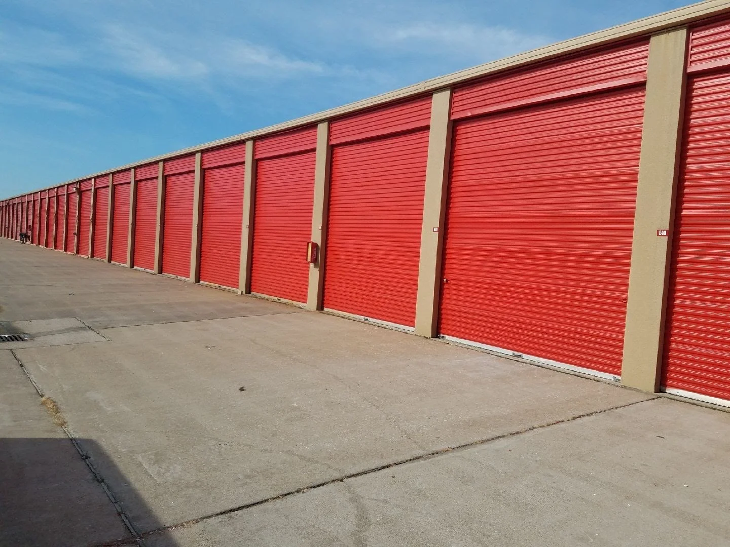 Row of industrial storage unit roll-up doors painted red, with a concrete sidewalk and blue sky in the background.
