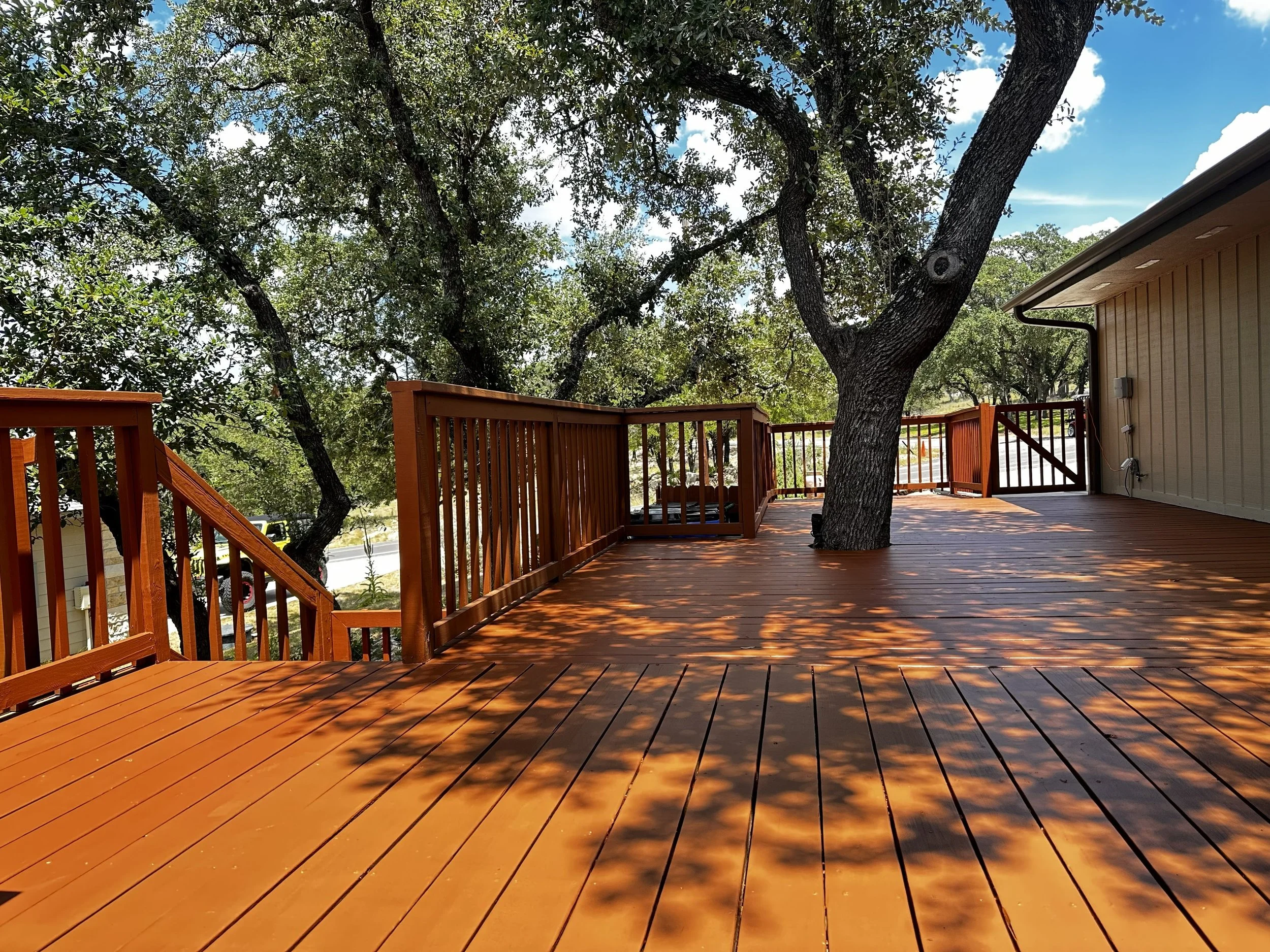 A wooden deck with a tree growing through it, casting shadows on the surface. To the right, part of a house with siding and a gutter. The sky is partly cloudy with blue sky visible.