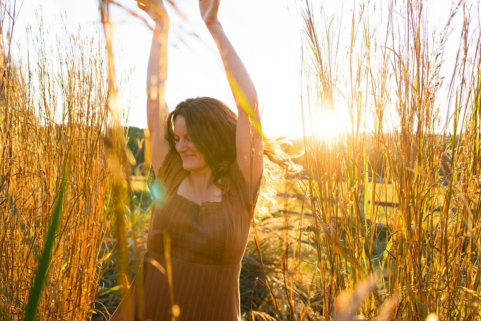 A woman in a brown dress standing in a golden wheat field at sunset, with her arms raised and eyes closed.