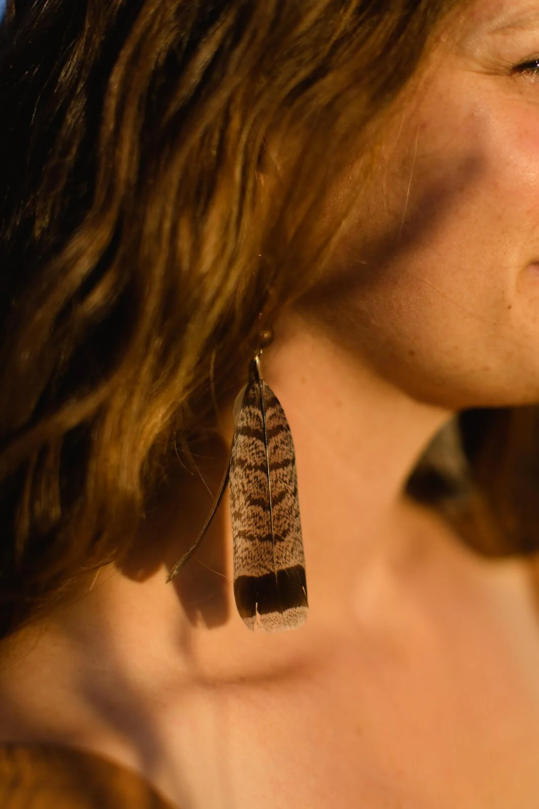 Close-up of a woman's face showing her ear and neck, with a large feather earring hanging from her ear, and her wavy brown hair partially covering her face.