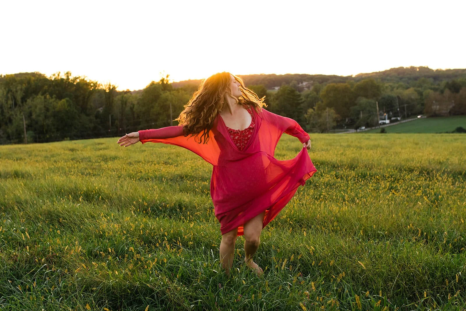 A woman with long, curly red hair dancing in a grassy field during sunset, wearing a flowing red dress.
