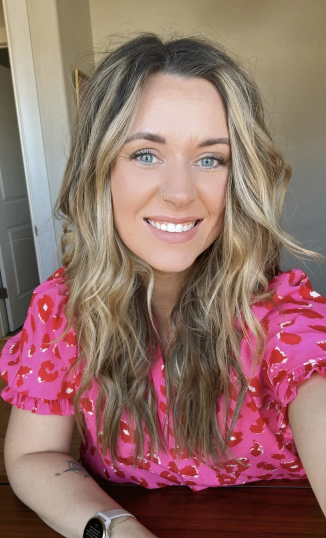 A woman with long wavy blonde hair and blue eyes smiling at the camera, wearing a pink dress with red heart patterns and a white watch, sitting at a wooden table inside a room.