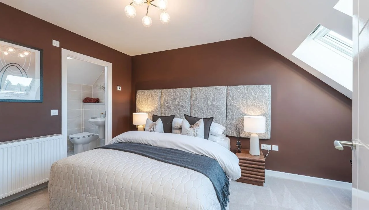 A bedroom with a bed, decorative pillows, and two bedside lamps. There is a small open bathroom with a toilet and sink. A skylight is on the sloped ceiling, and there is a framed picture on the wall.