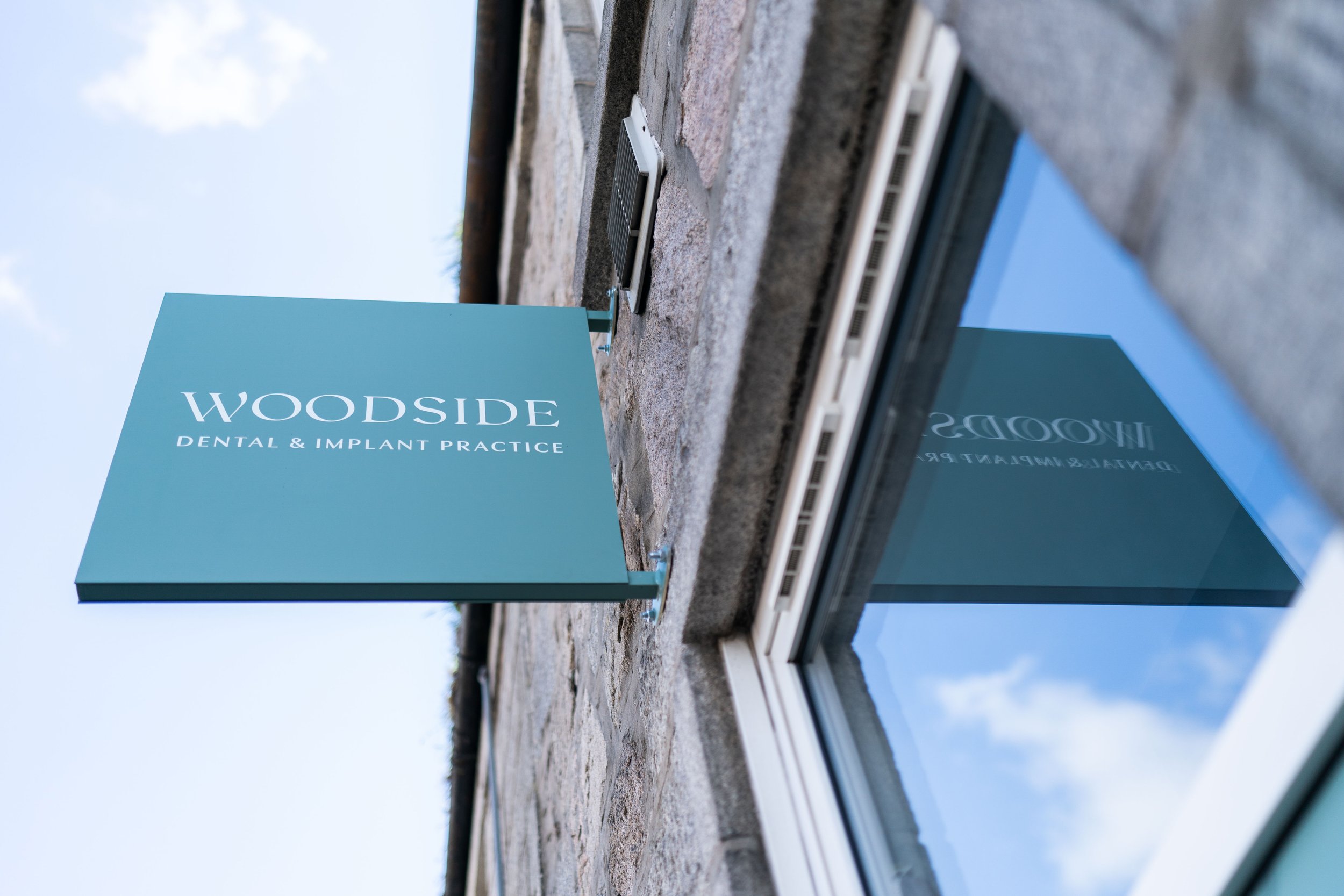 Sign for Woodsid Dental & Implant Practice attached to a building exterior with a stone facade, under a clear blue sky.