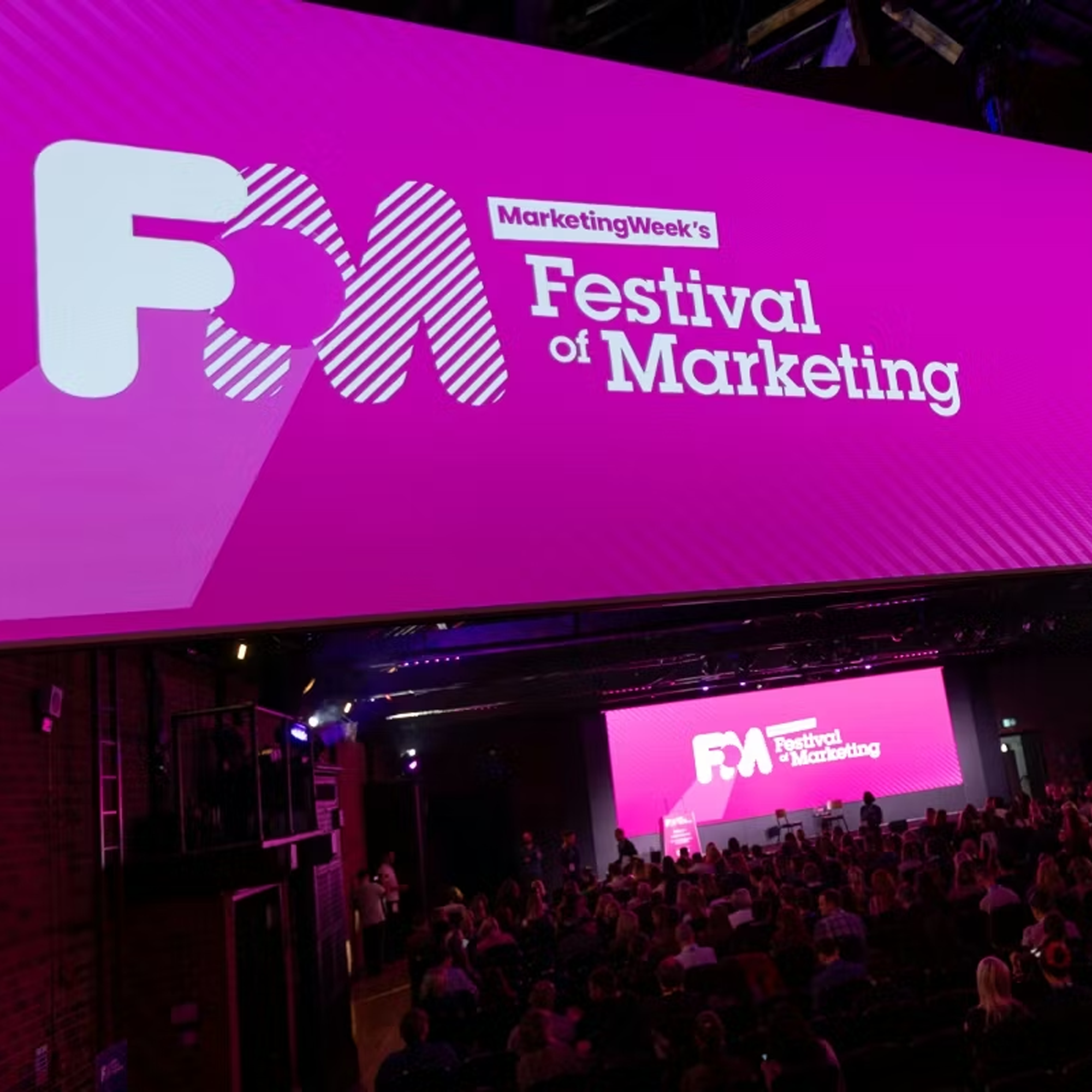 A large conference room filled with attendees at the Festival of Marketing event. The stage has a large pink screen displaying the event logo, with the words "Marketing Week's Festival of Marketing." The room is dimly lit with purple and pink lighting, and people are sitting and standing while listening to a speaker.
