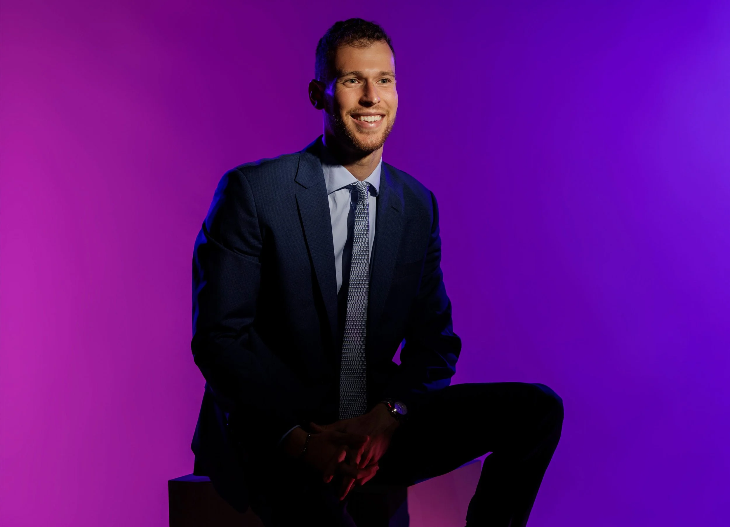 A man in a dark suit with a light-colored shirt and patterned tie, smiling, posed against a vibrant purple background.