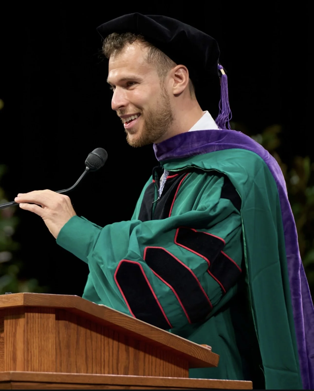 A young man wearing a graduation cap and gown with purple, green, and black colors, speaking at a podium with a microphone.